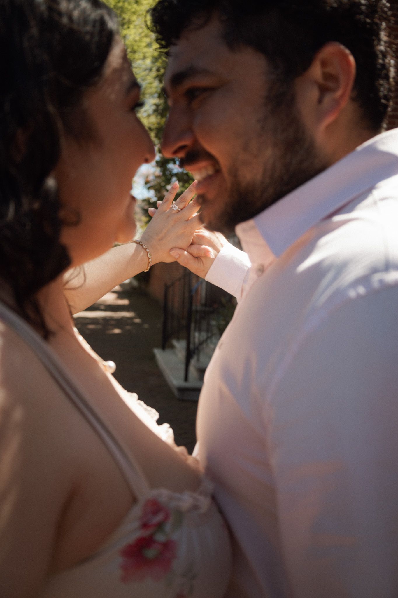 Couple sharing a romantic moment outdoors, woman touching man's face, both smiling, wearing casual attire in sunlight.