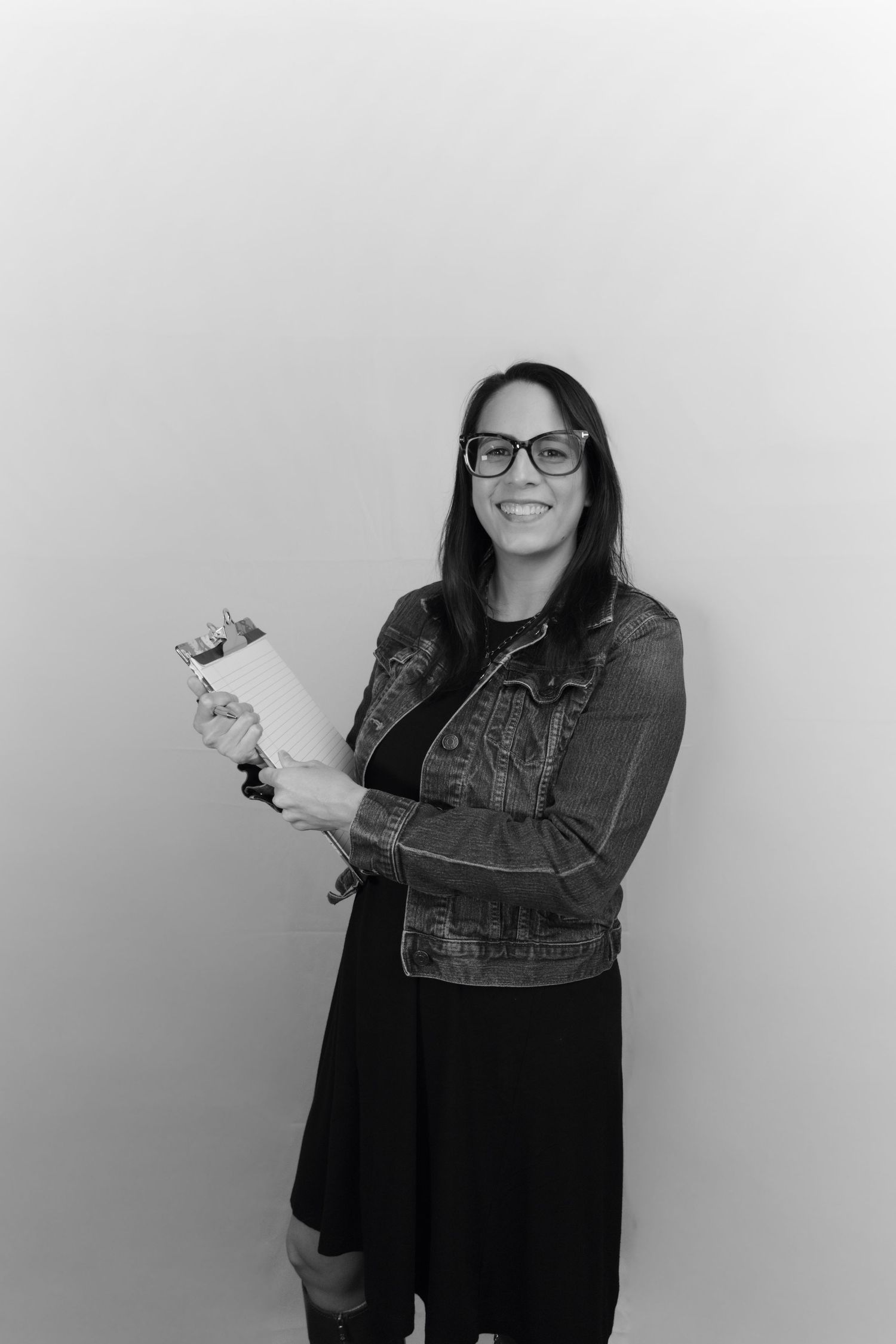 Black and white photo of a smiling woman in glasses and denim jacket holding a clipboard against a light background.