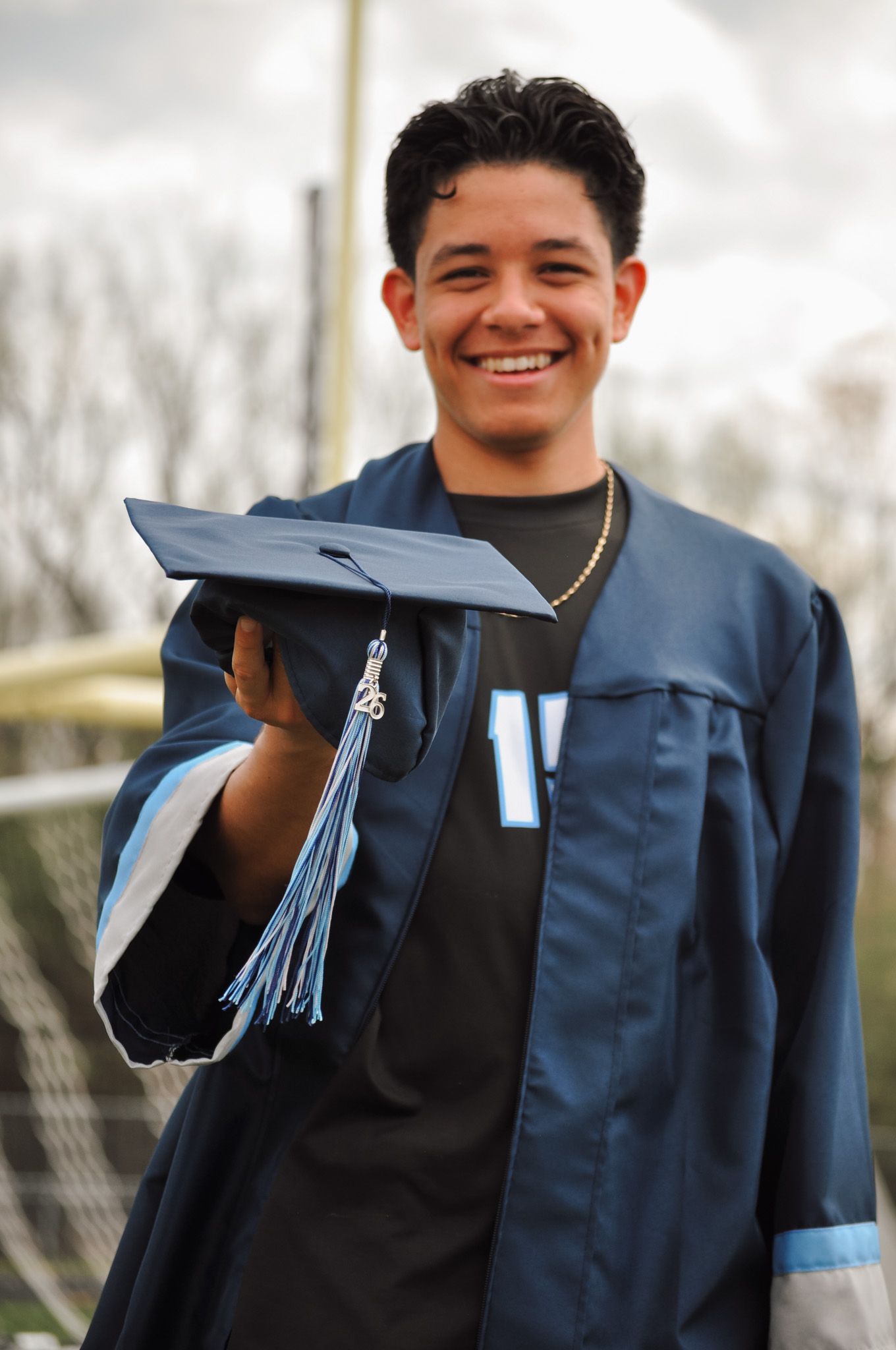 Smiling young man in navy blue graduation gown holding cap with tassel outdoors.