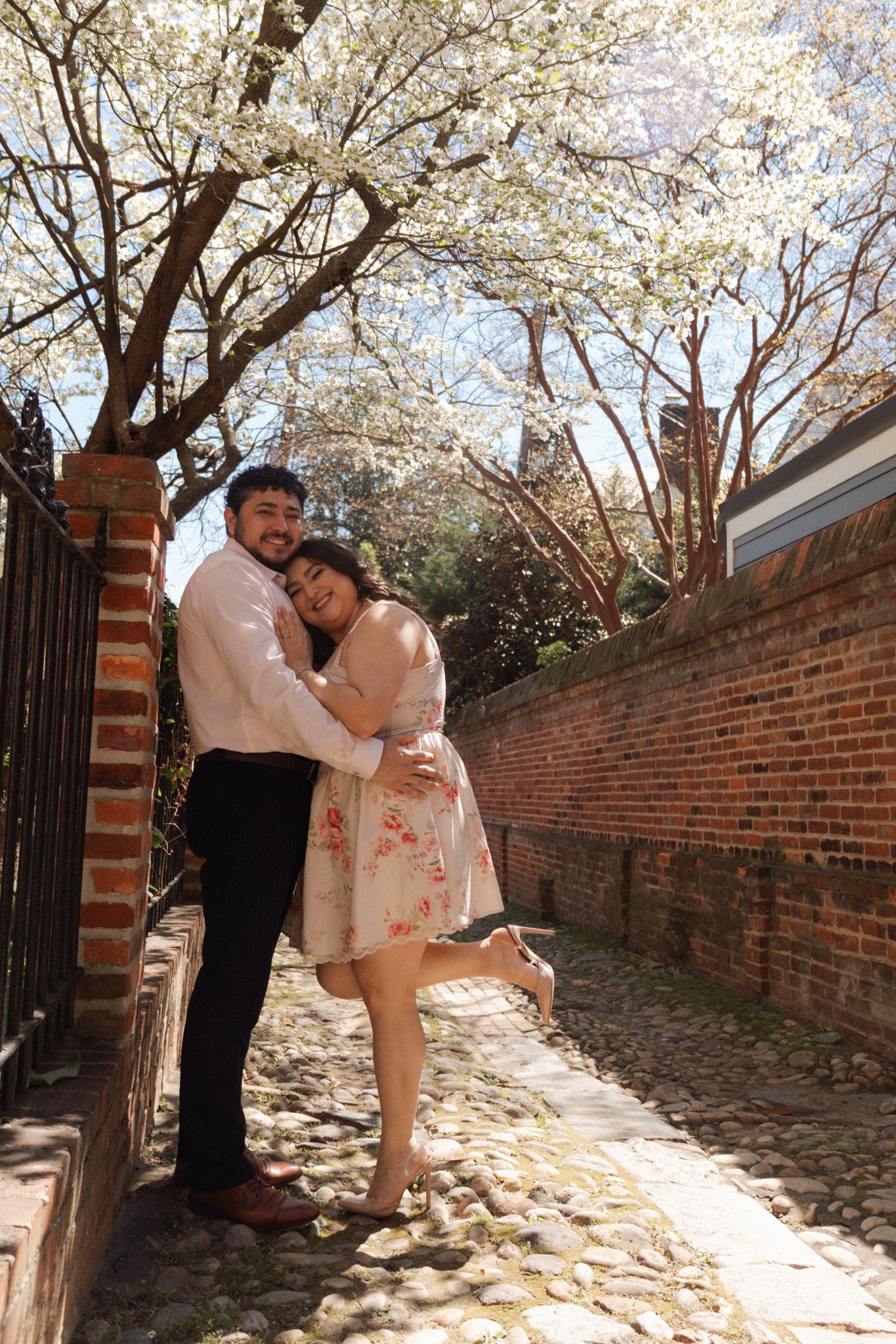 Couple embracing and smiling under blooming white cherry blossom trees on a cobblestone path beside a brick wall.