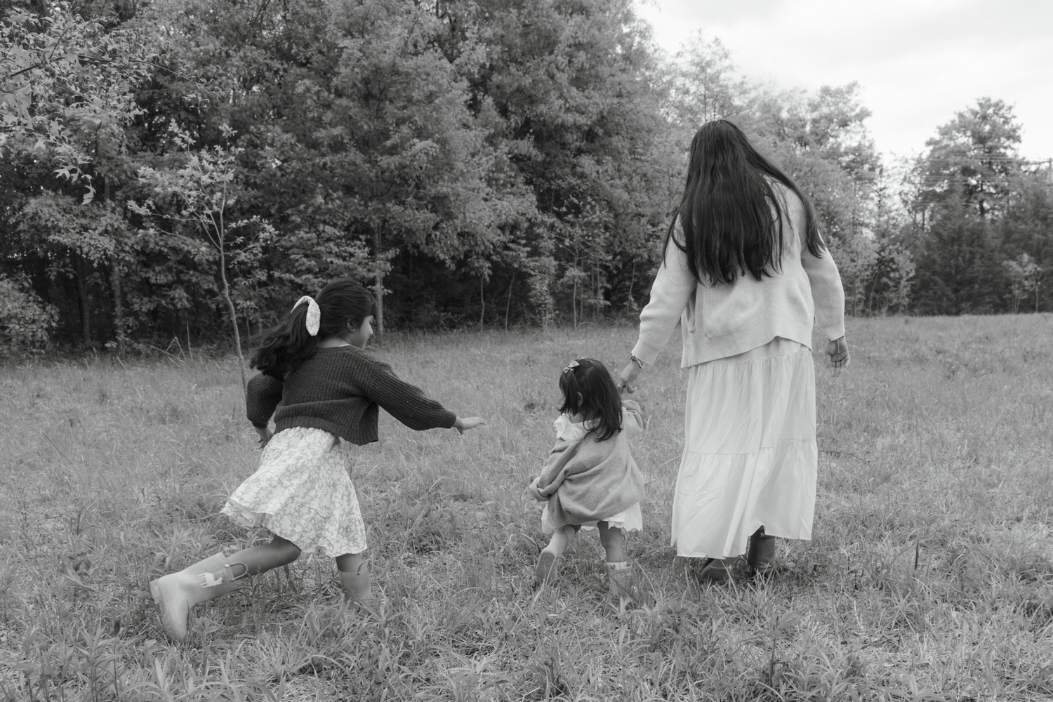 Black and white photo of two children and a woman in a white dress walking through a grassy field near trees.