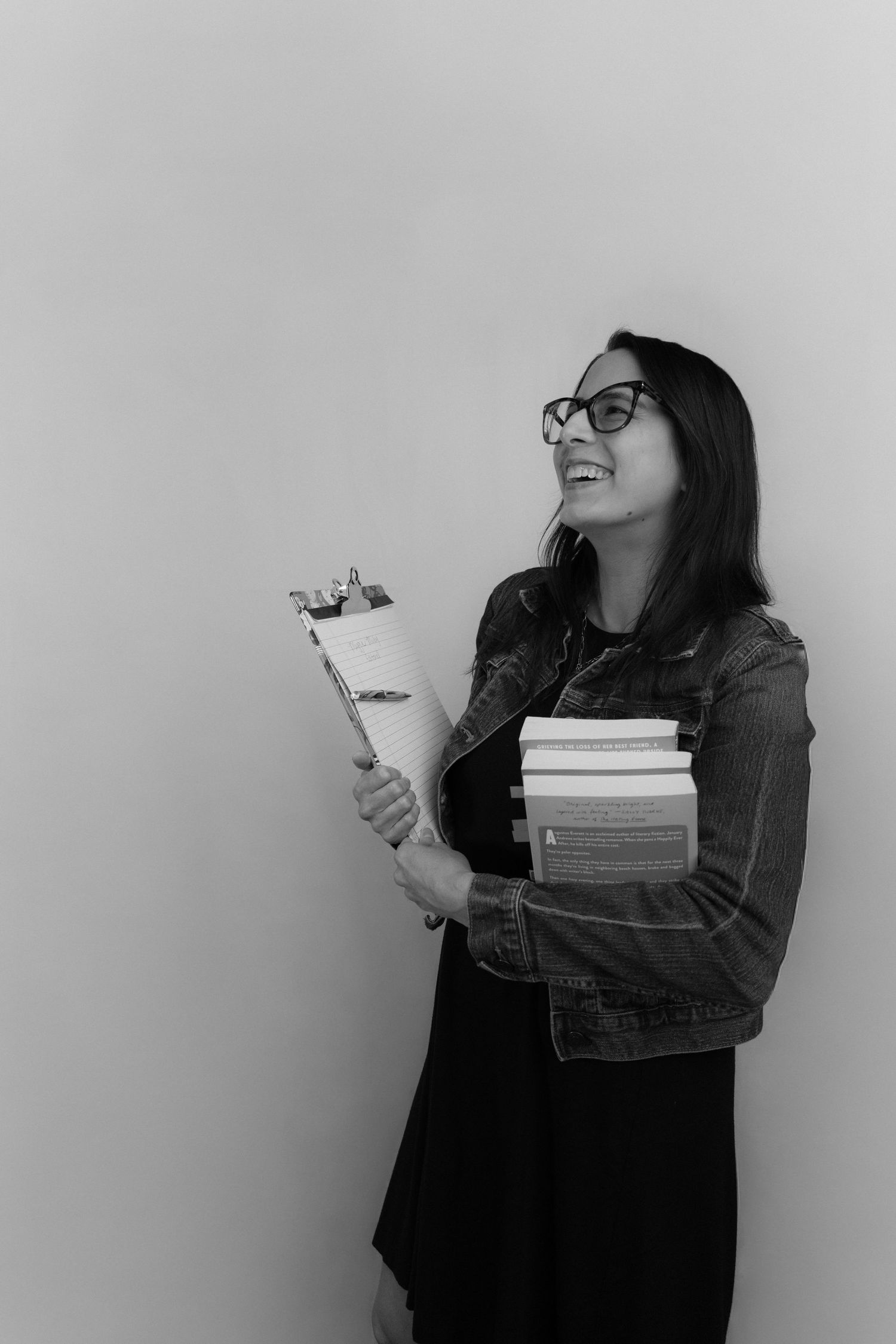 Smiling woman with glasses holding clipboard and stack of books in black and white photo.