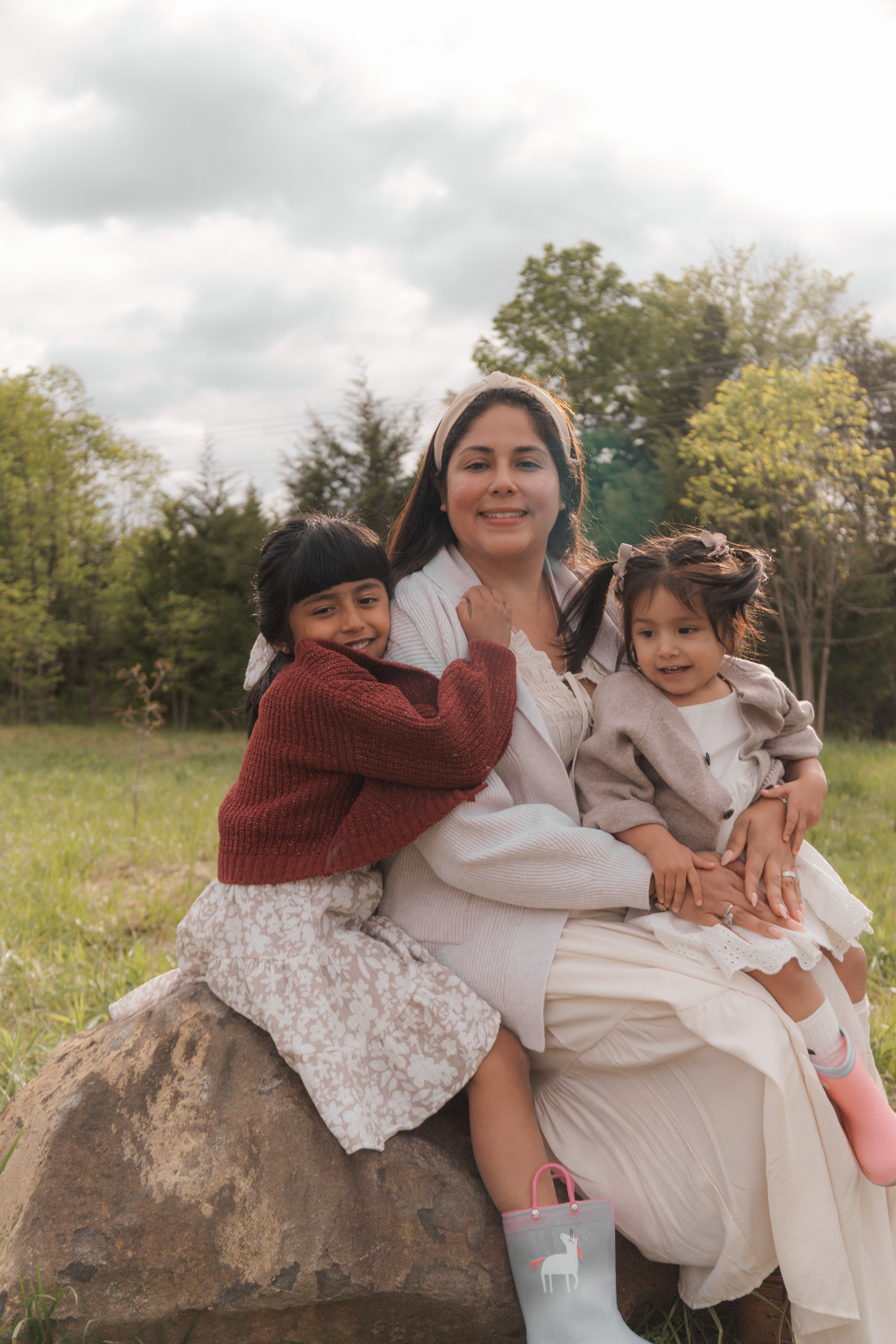 A smiling woman sits on a rock outdoors with two young girls hugging her, surrounded by green trees.