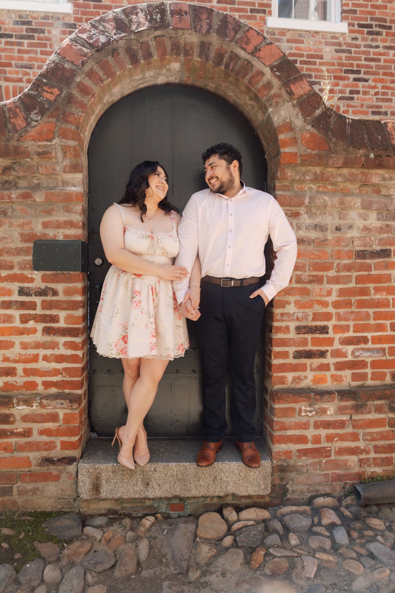 Couple holding hands and smiling in front of a rustic brick archway door, dressed in elegant casual attire.