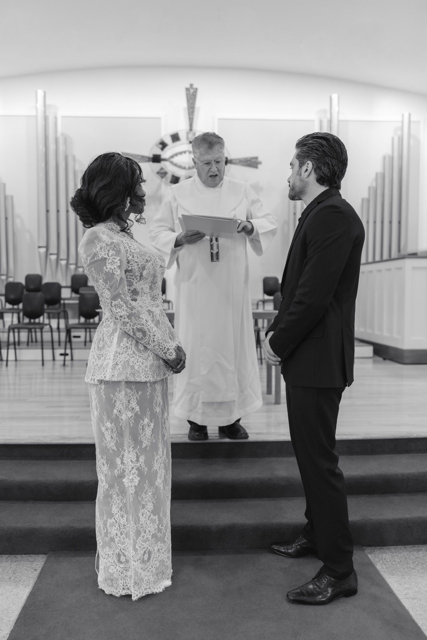 Black and white photo of a couple exchanging wedding vows before an officiant in a modern church ceremony.