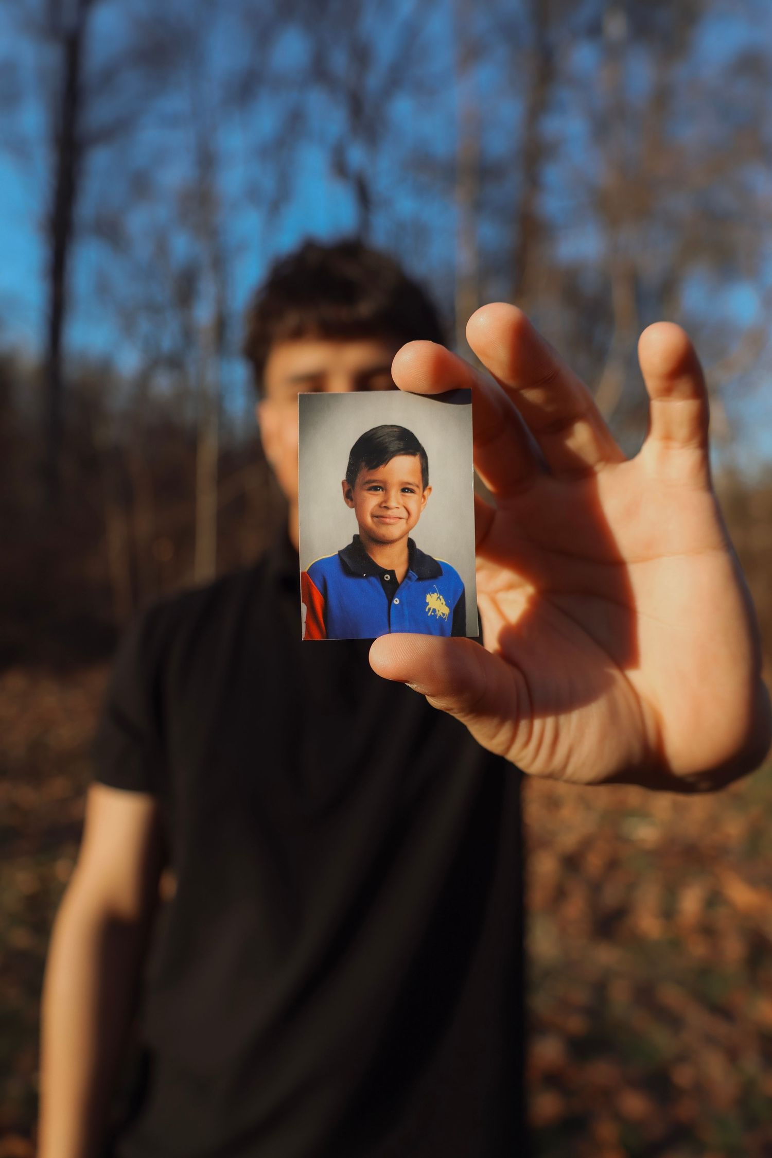 A teenager holds up a childhood school photo portrait outdoors in a forest setting with blue sky.