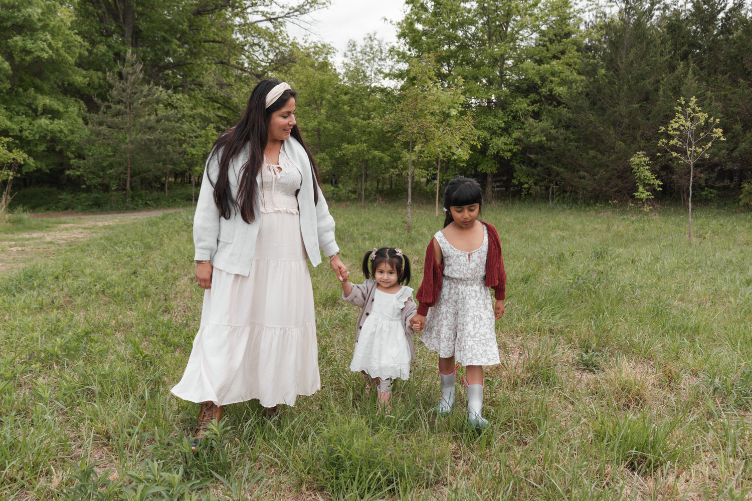 A mother and two young daughters walk hand-in-hand through a green meadow, wearing matching white and floral dresses.