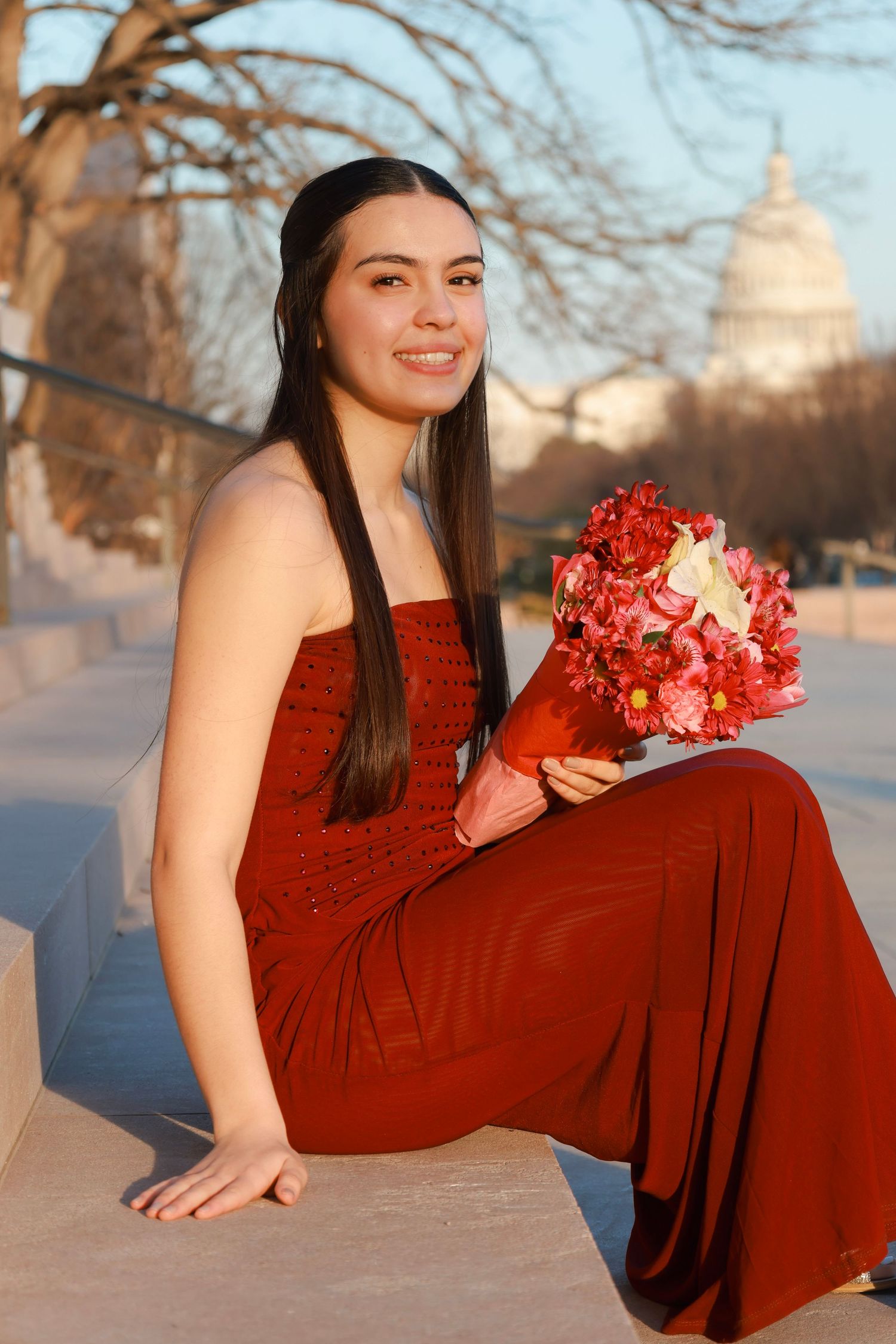 Young woman in red dress holding pink bouquet sits on steps near US Capitol building at golden hour.