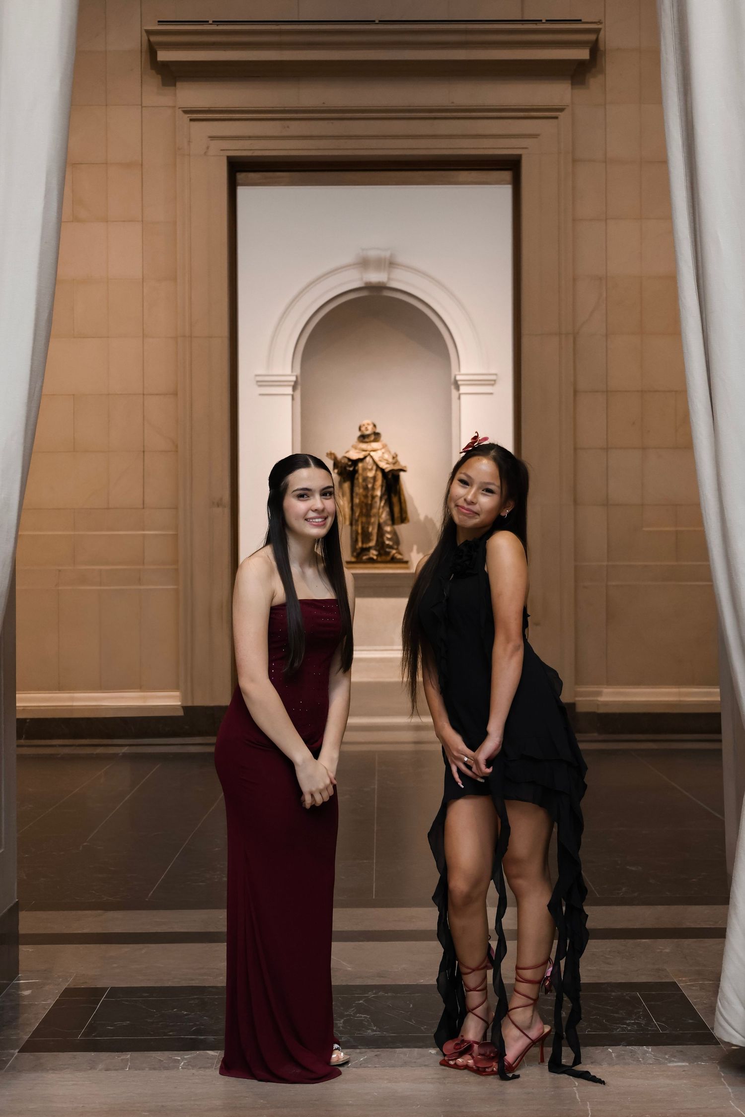 Two young women in elegant black and burgundy formal gowns posing in a grand hall with ornate architectural details.