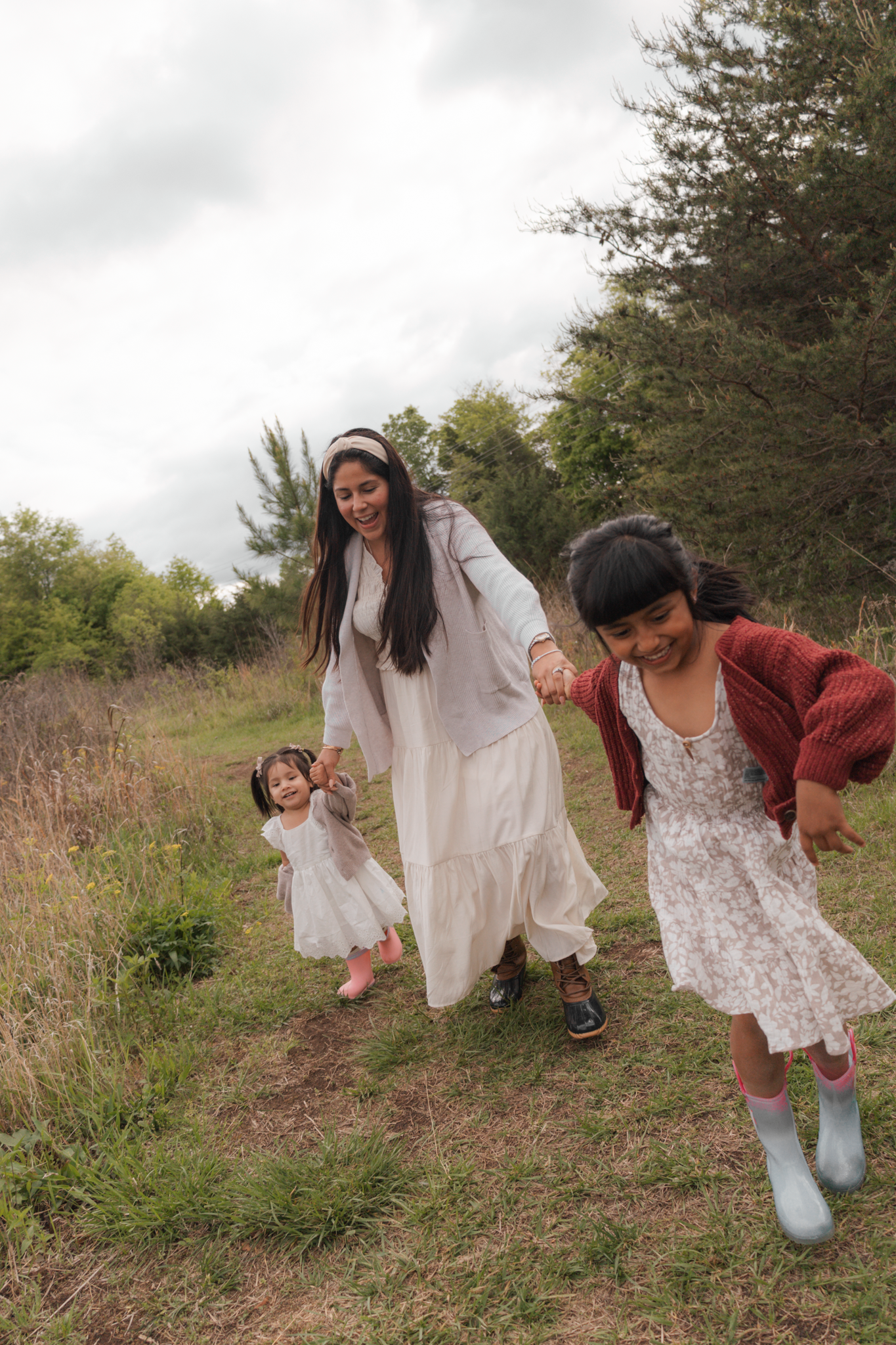A mother and two young daughters walk hand-in-hand through a grassy field surrounded by trees on a cloudy day.