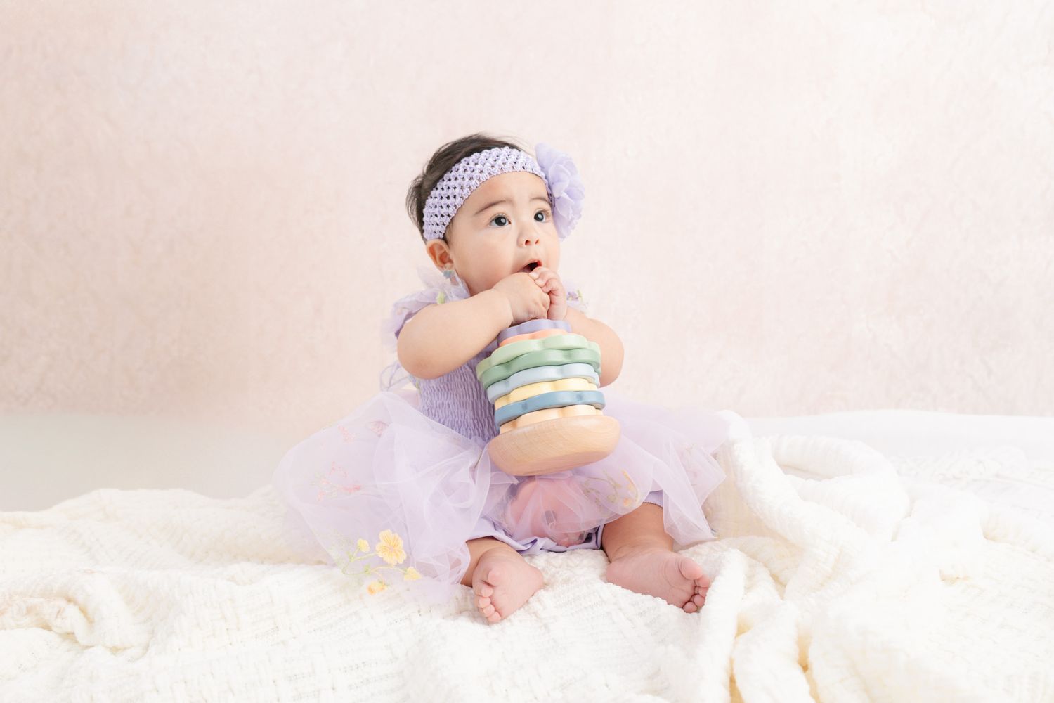 A cute baby girl in a purple tutu dress and headband sits on a white blanket, chewing on a colorful stacking toy.