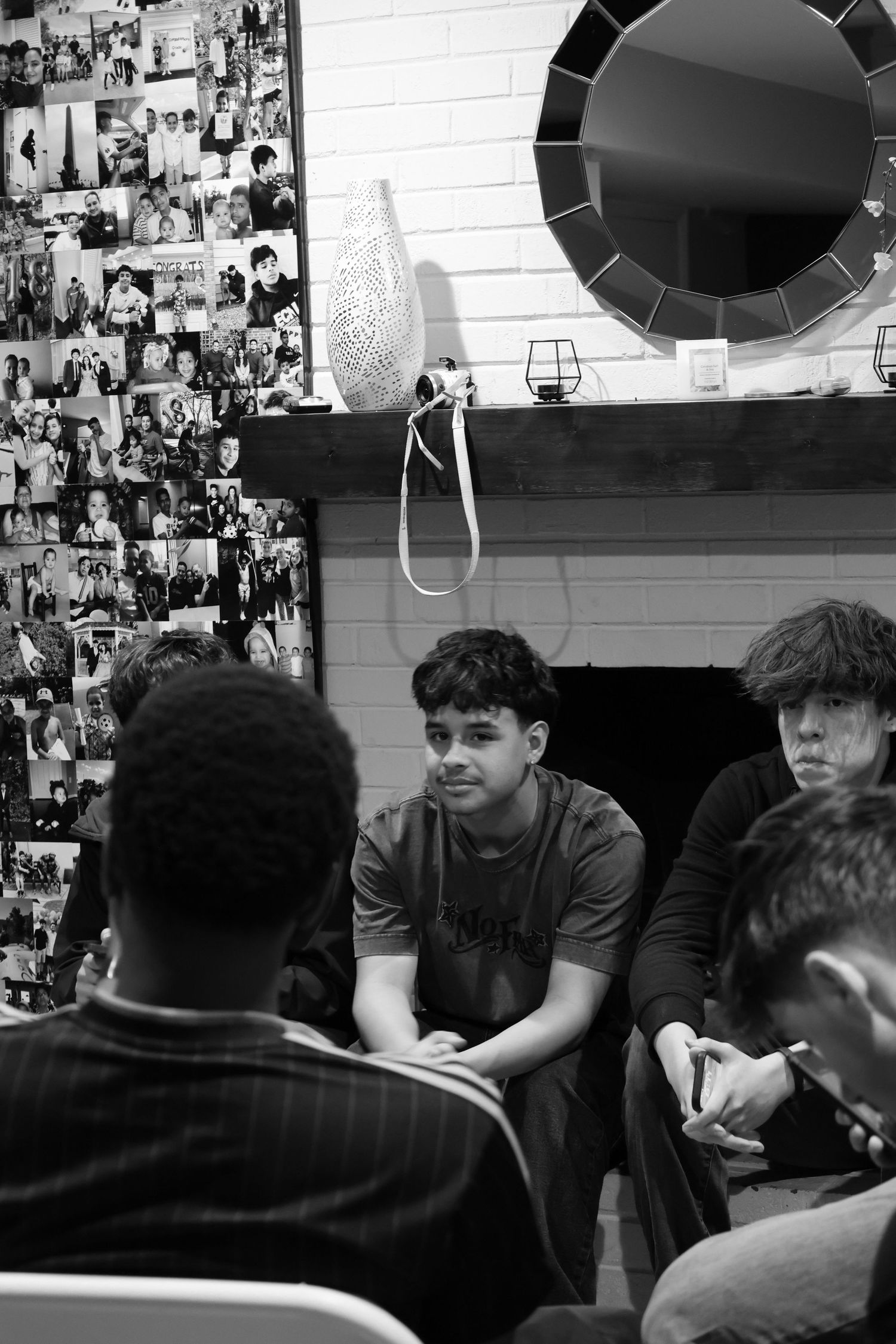Black and white photo of young men gathered around a table in a room decorated with photo collages on the wall.