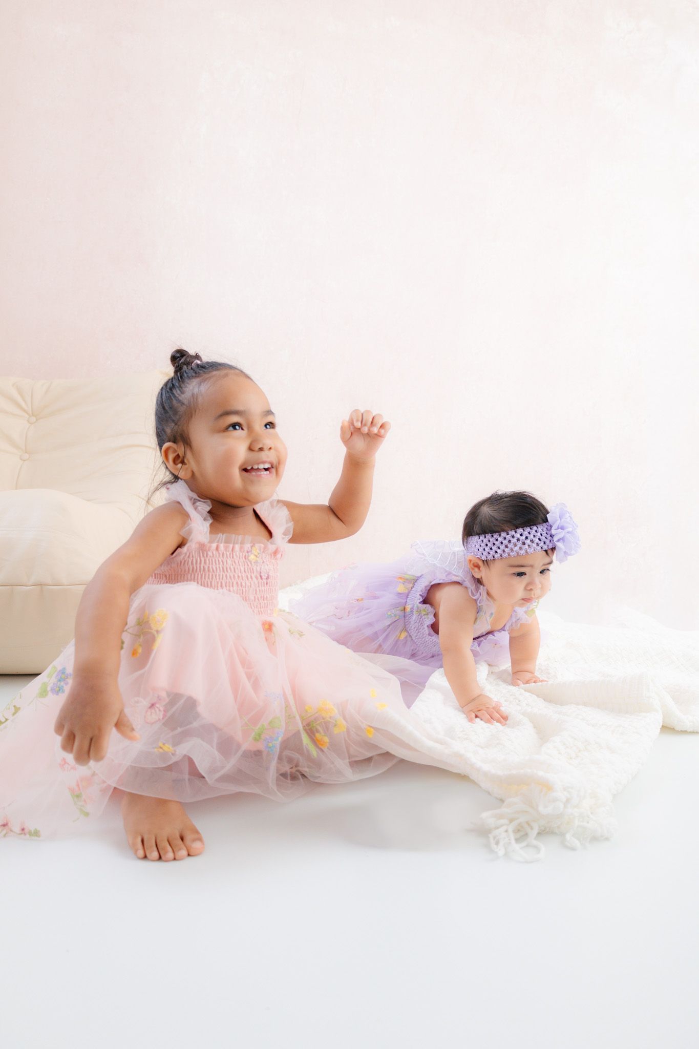 Two young girls in tulle dresses play on a white floor, one in pink smiling brightly, the other in lavender with a floral headband.