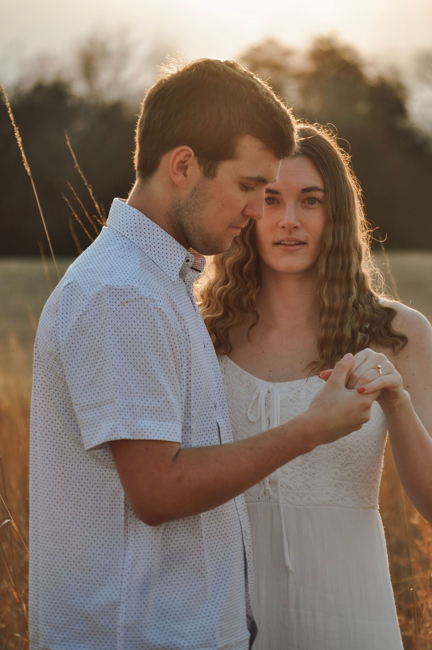 A couple shares a tender moment in a golden sunlit field, the man kissing the woman's forehead as she gazes softly ahead.
