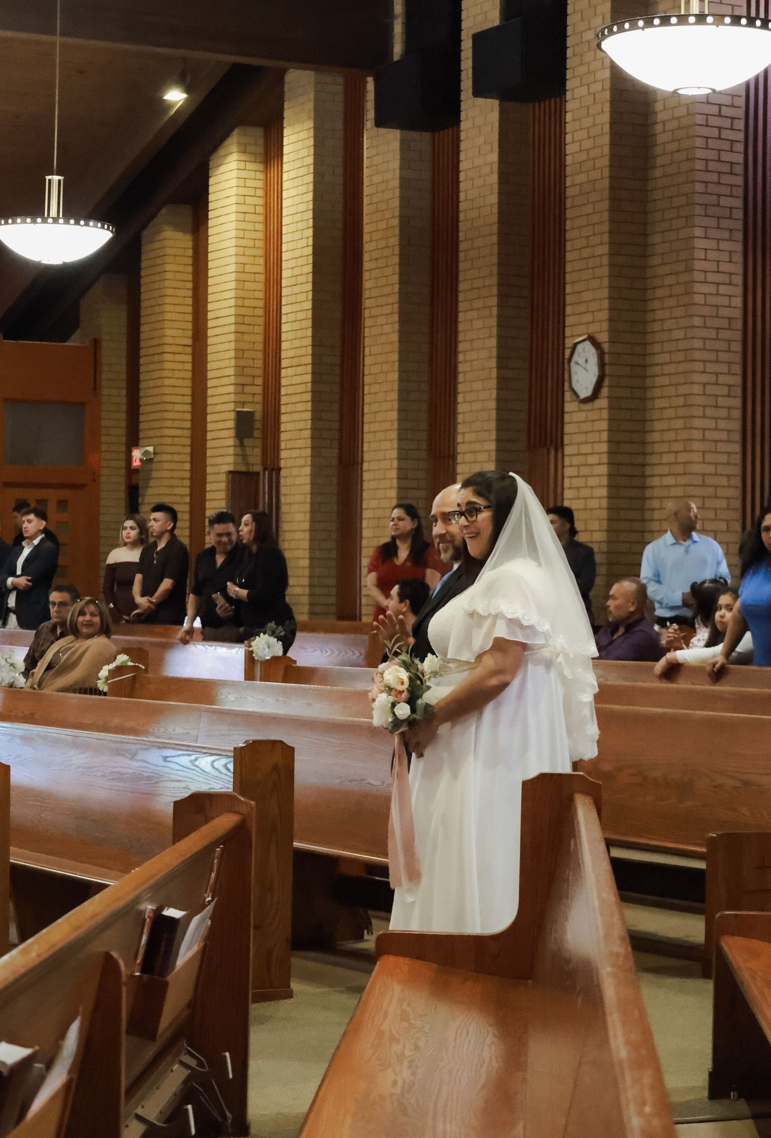 Bride and groom in white dress and veil walks down church aisle holding bouquet, with guests seated in wooden pews.