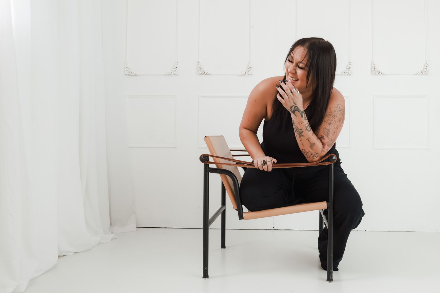A tattooed woman in a black dress sits thoughtfully on a modern chair in a bright white studio setting.