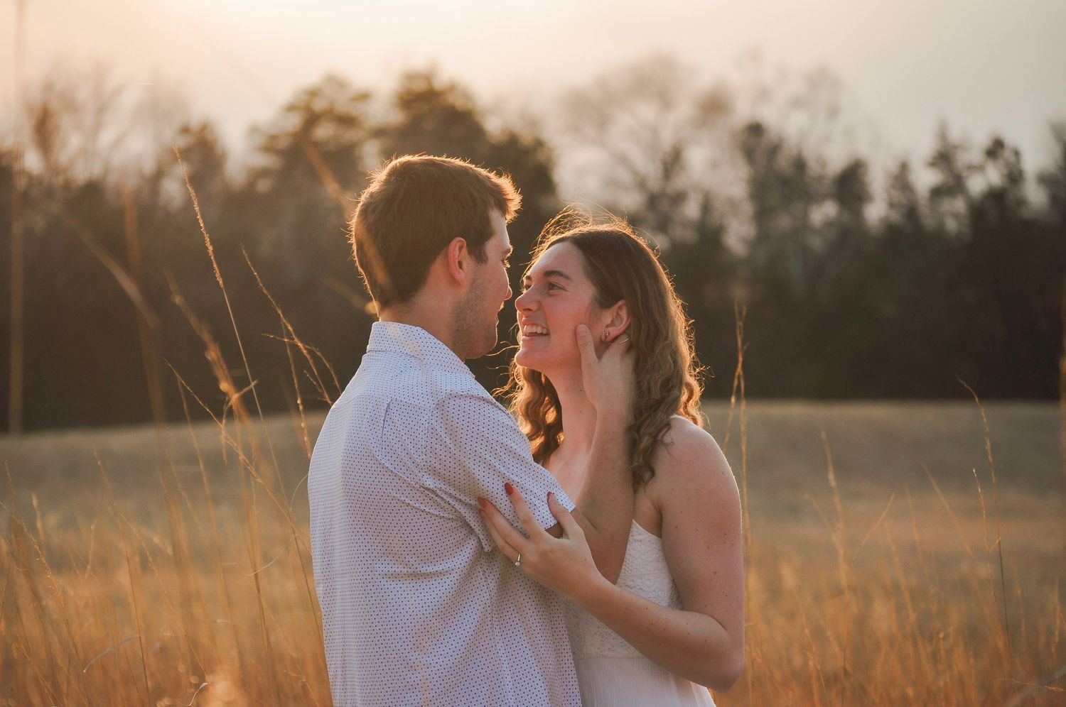 Couple embracing in golden sunset field, woman smiling up at man, tall grass and trees in warm backlight.