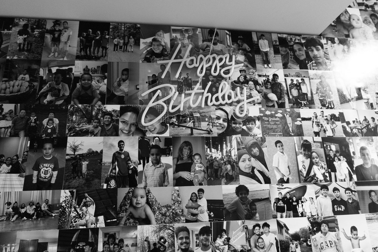 A wall covered with black and white photos collaged together, featuring a neon Happy Birthday sign centerpiece.