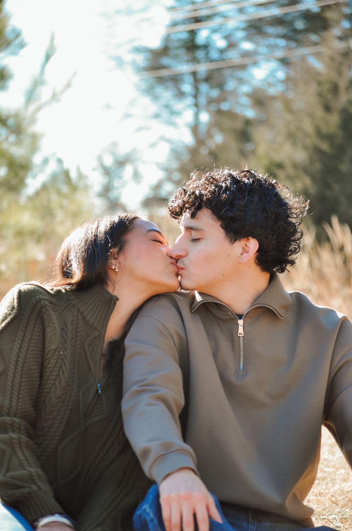 A young couple shares a romantic kiss outdoors, sitting together in a sunny natural setting.