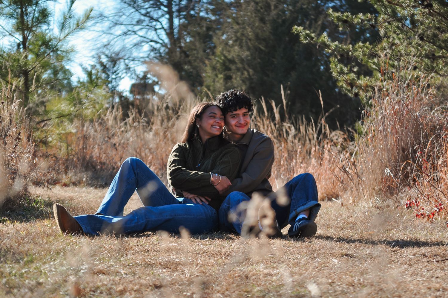 Young couple sitting together in a sunny field with dry grass and trees, sharing a romantic outdoor moment with a small dog.
