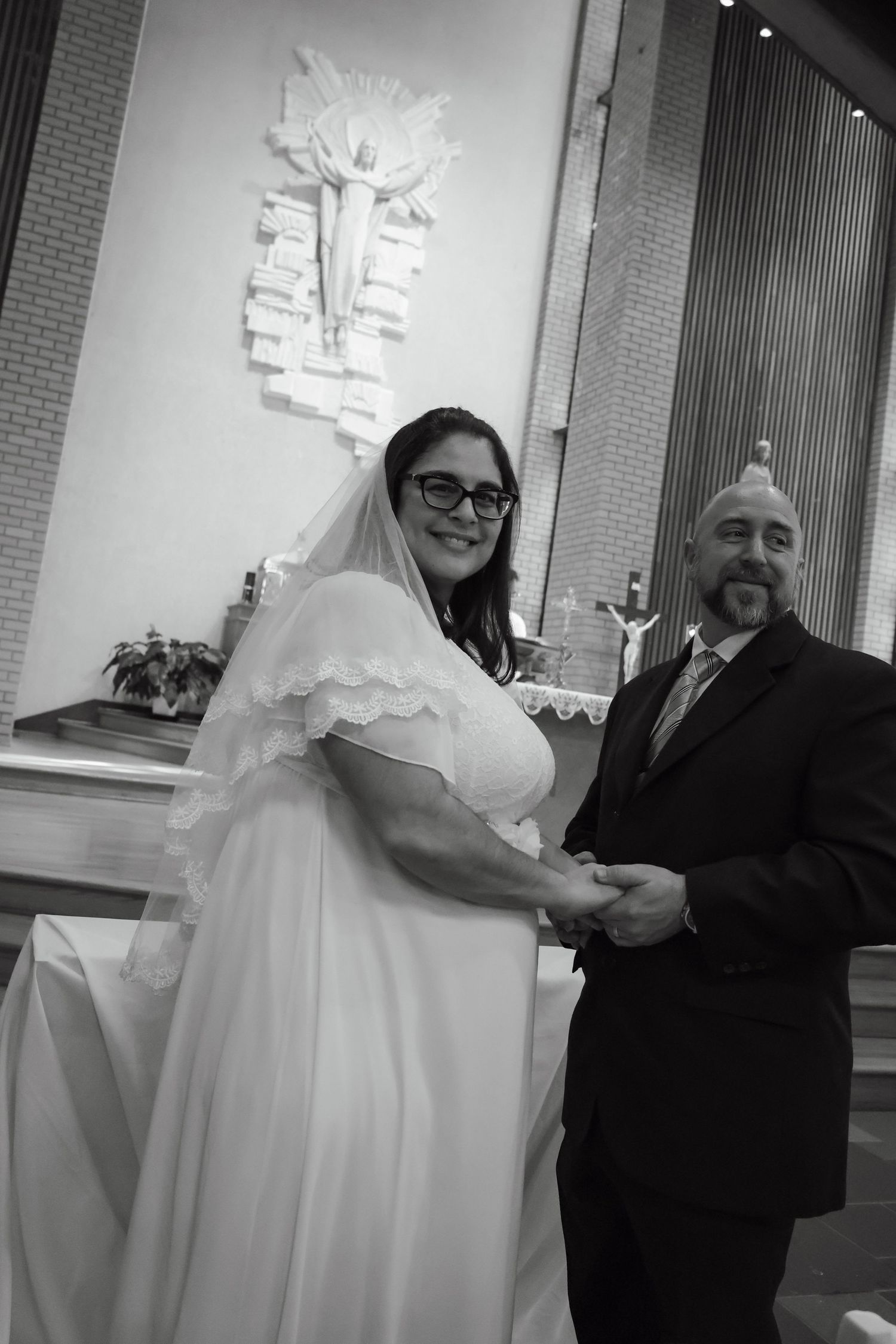 Black and white photo of a smiling bride in white gown and veil holding hands with groom in suit at church altar.