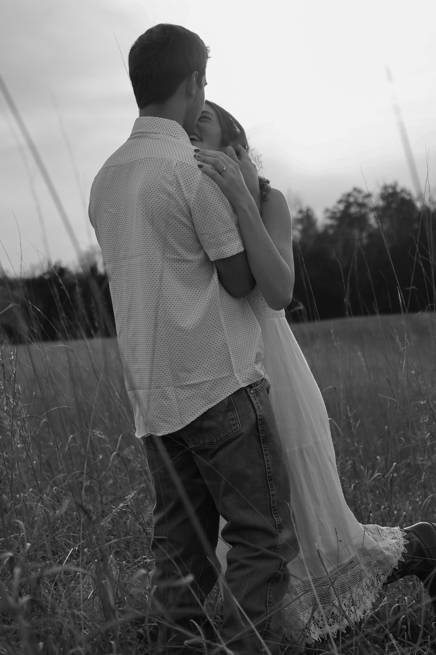 Black and white photo of a couple embracing romantically in a field at dusk, woman in white dress leaning into mans arms.