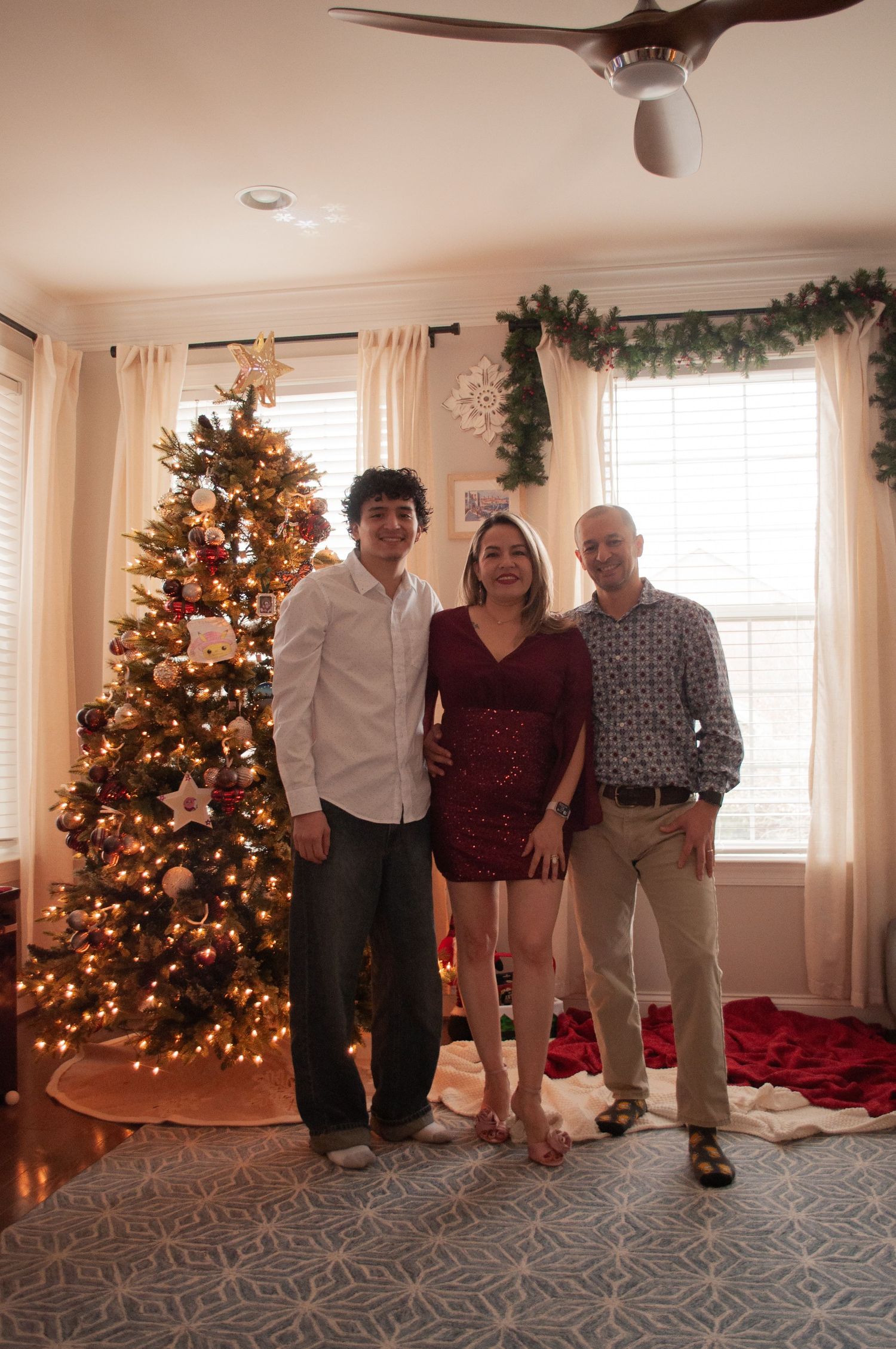 Three people posing together in front of a decorated Christmas tree in a festive living room with holiday garland.