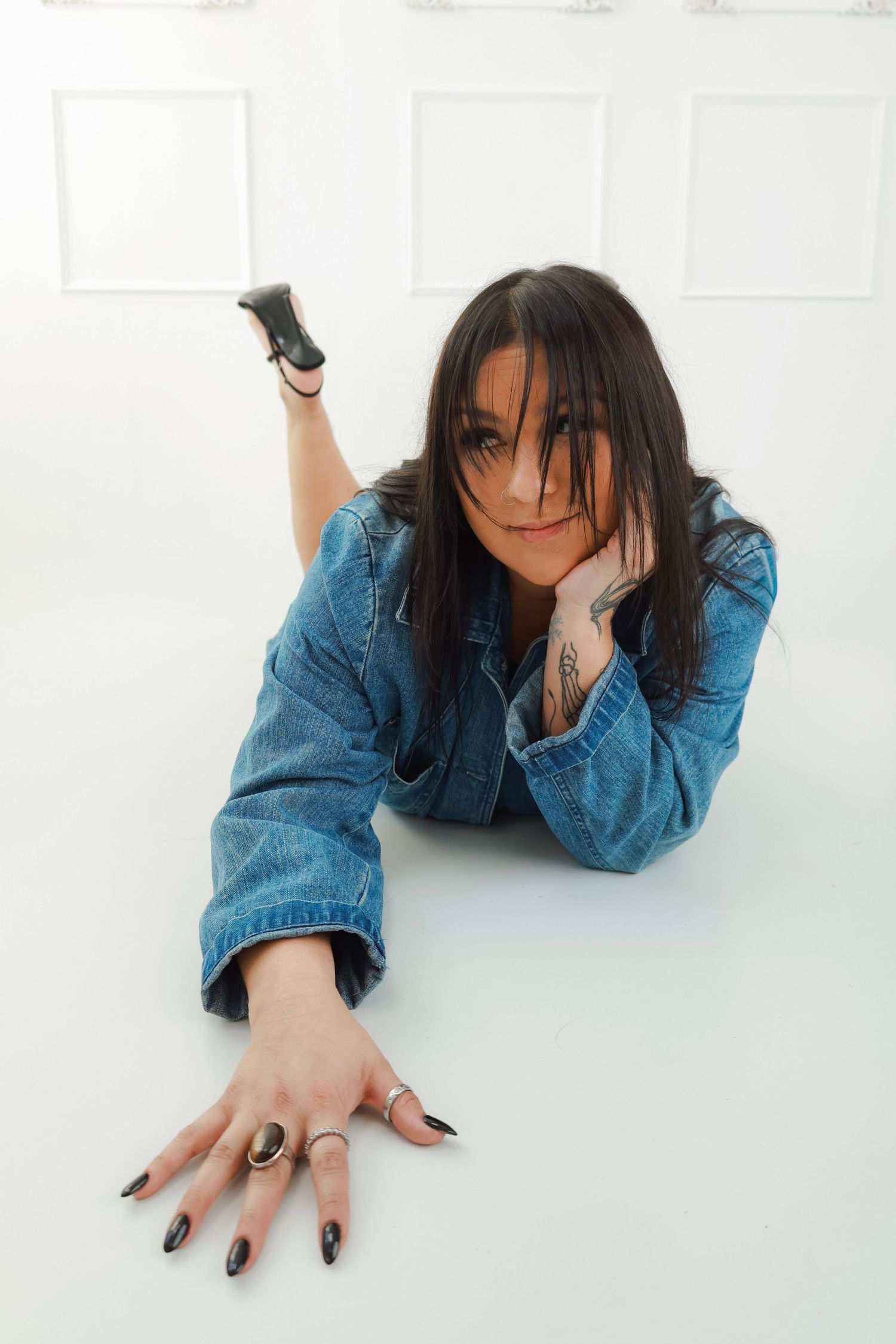 Woman in denim jacket lying on white floor, showing tattoos and black nail polish, with rings on fingers.