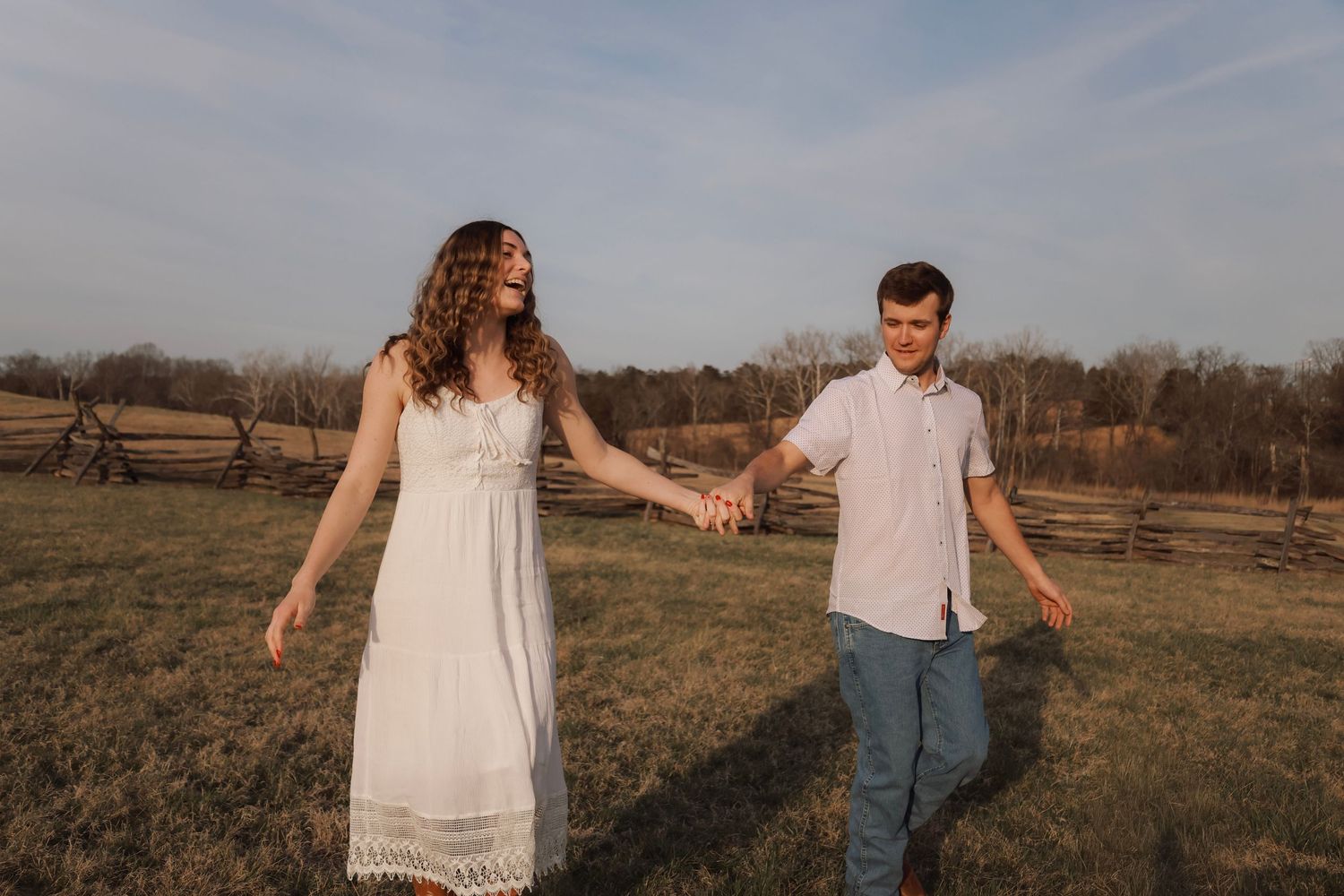 Couple holding hands walking in a golden field at sunset, woman in white dress, man in casual attire.