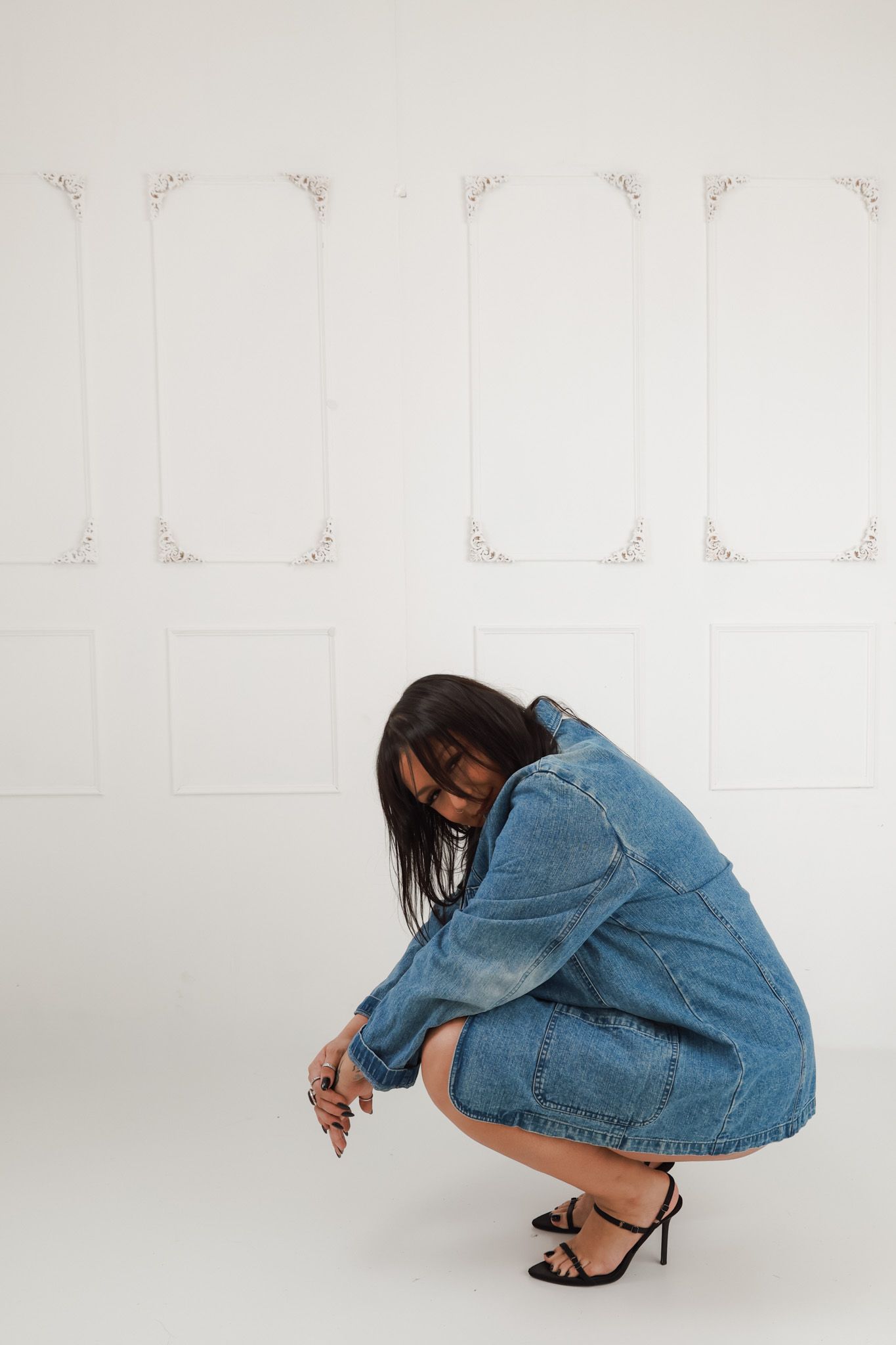 Woman in oversized denim dress crouching in heels against white decorative wall panel backdrop.