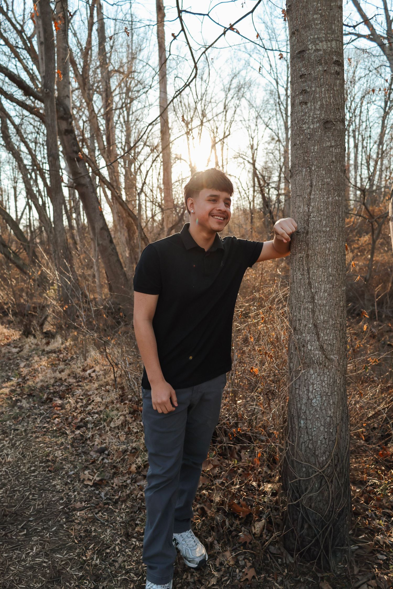 Young man in black shirt leaning against a tree in autumn woods with golden sunlight filtering through bare branches.