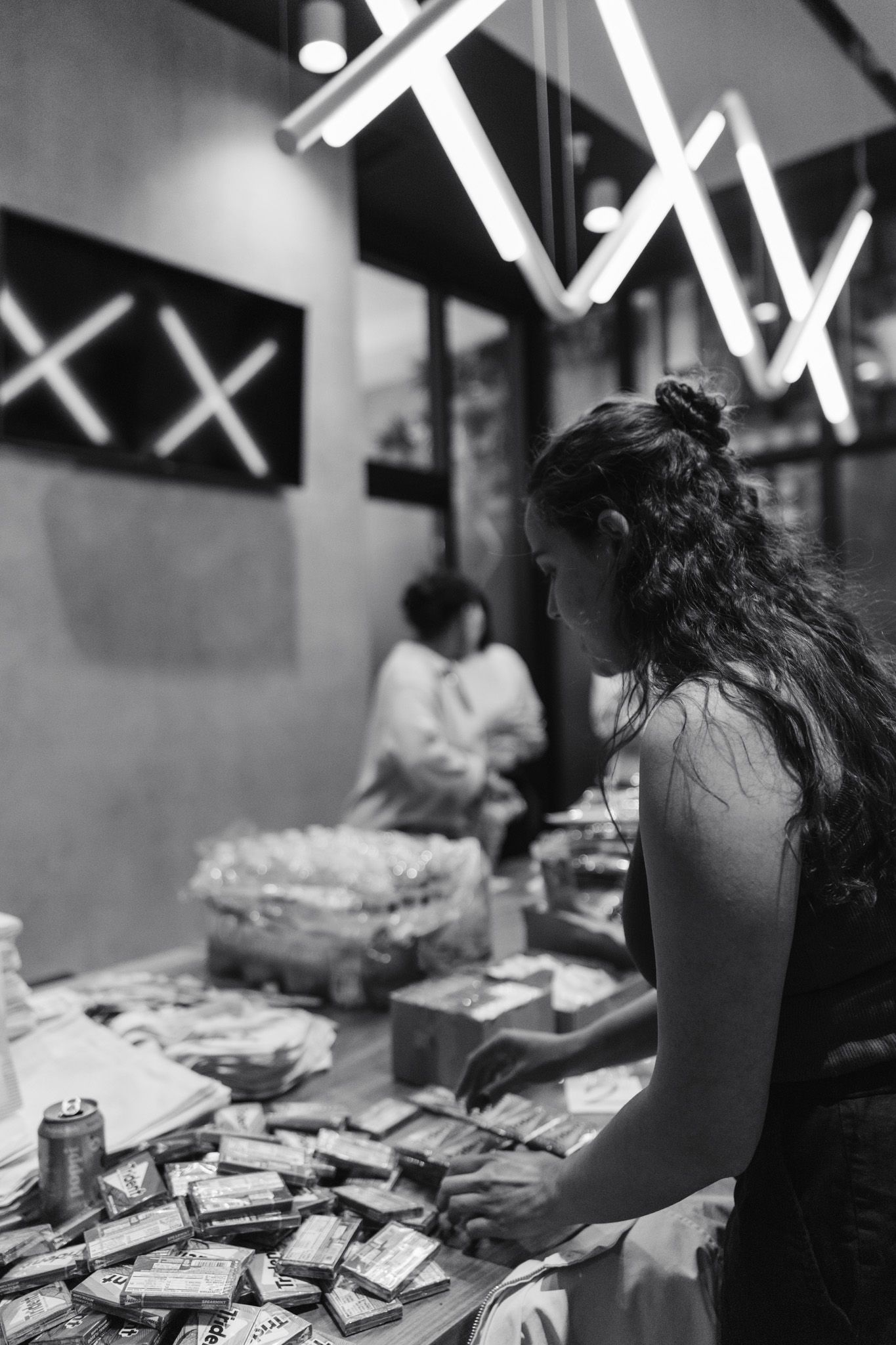 Black and white photo of a woman with curly hair sorting items on a table at an indoor market or pop-up shop.