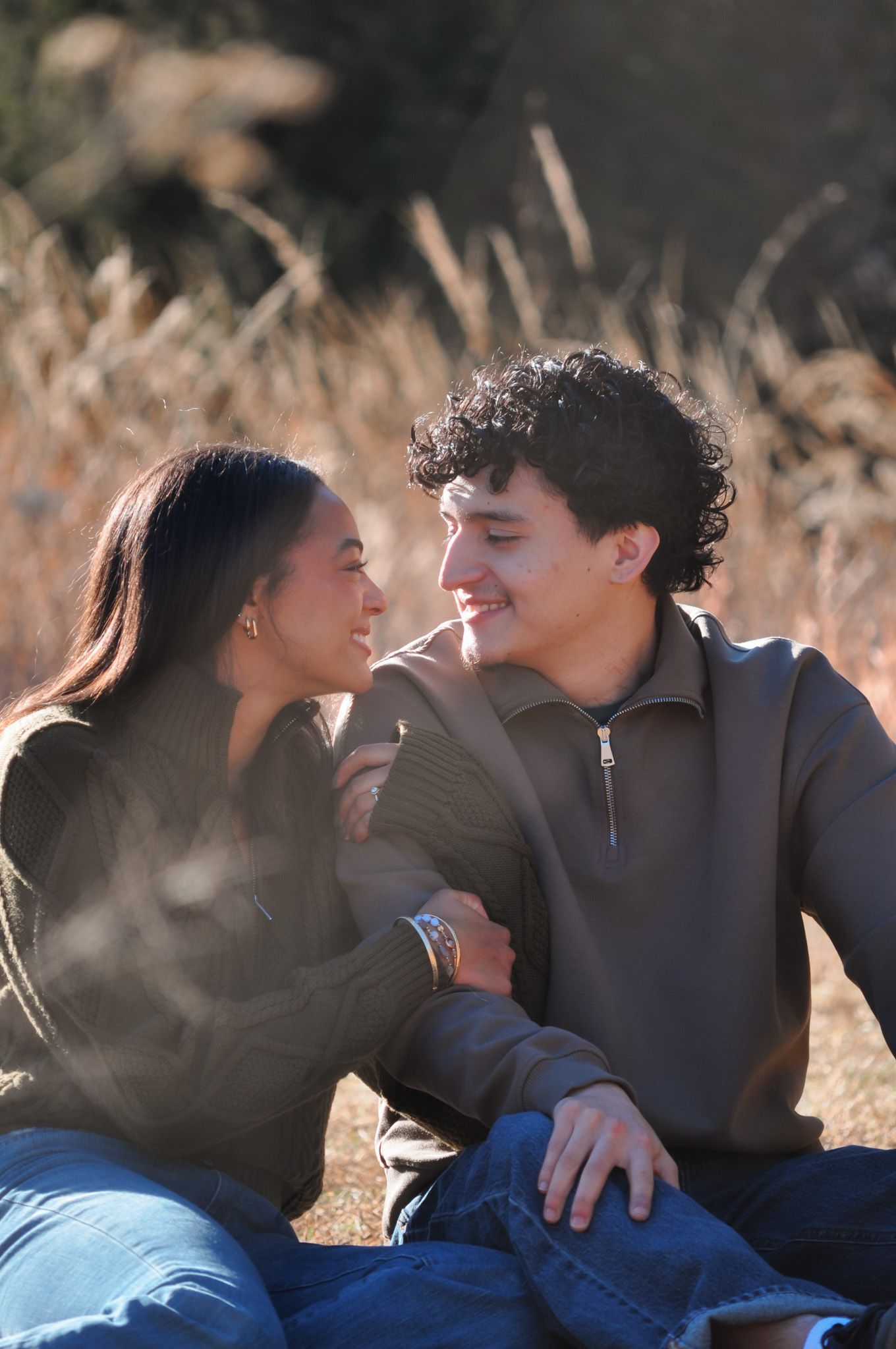 A young couple sits together in a golden field, smiling and gazing at each other in a warm, romantic outdoor setting.