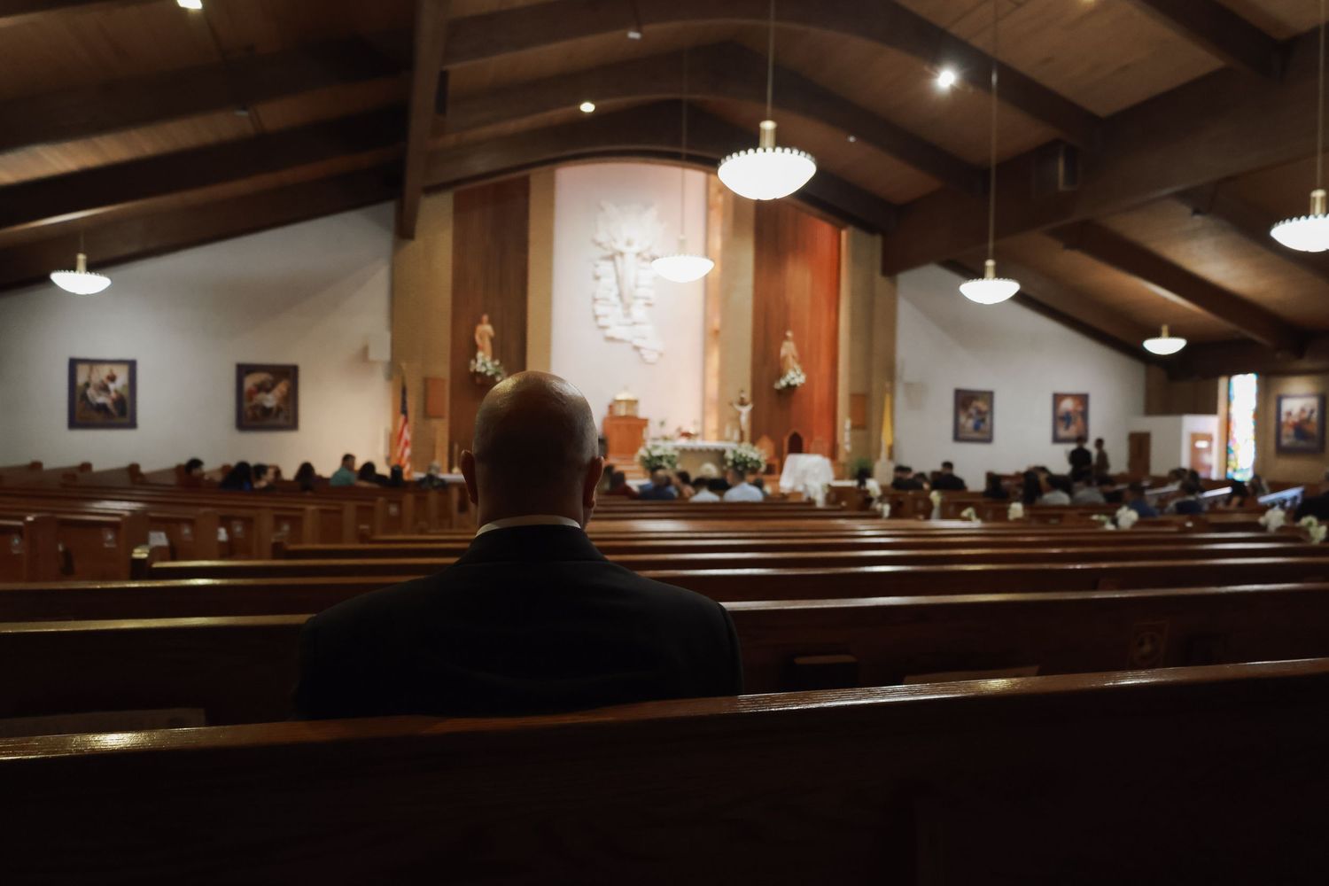 Man sitting alone in wooden church pew during a Catholic mass service with altar and religious artwork visible.