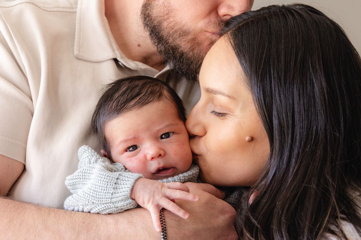 Parents kissing their newborn baby on the cheeks in a tender family portrait.