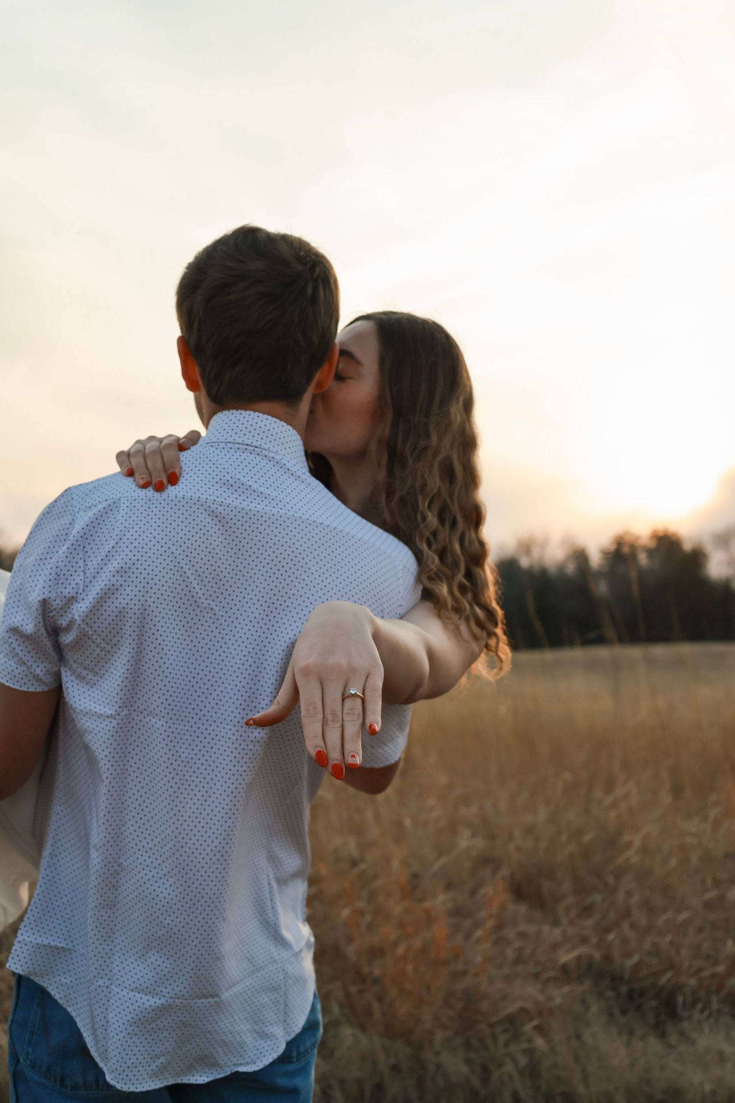 A couple embraces at sunset in a golden field, woman kissing mans cheek while showing off her engagement ring.
