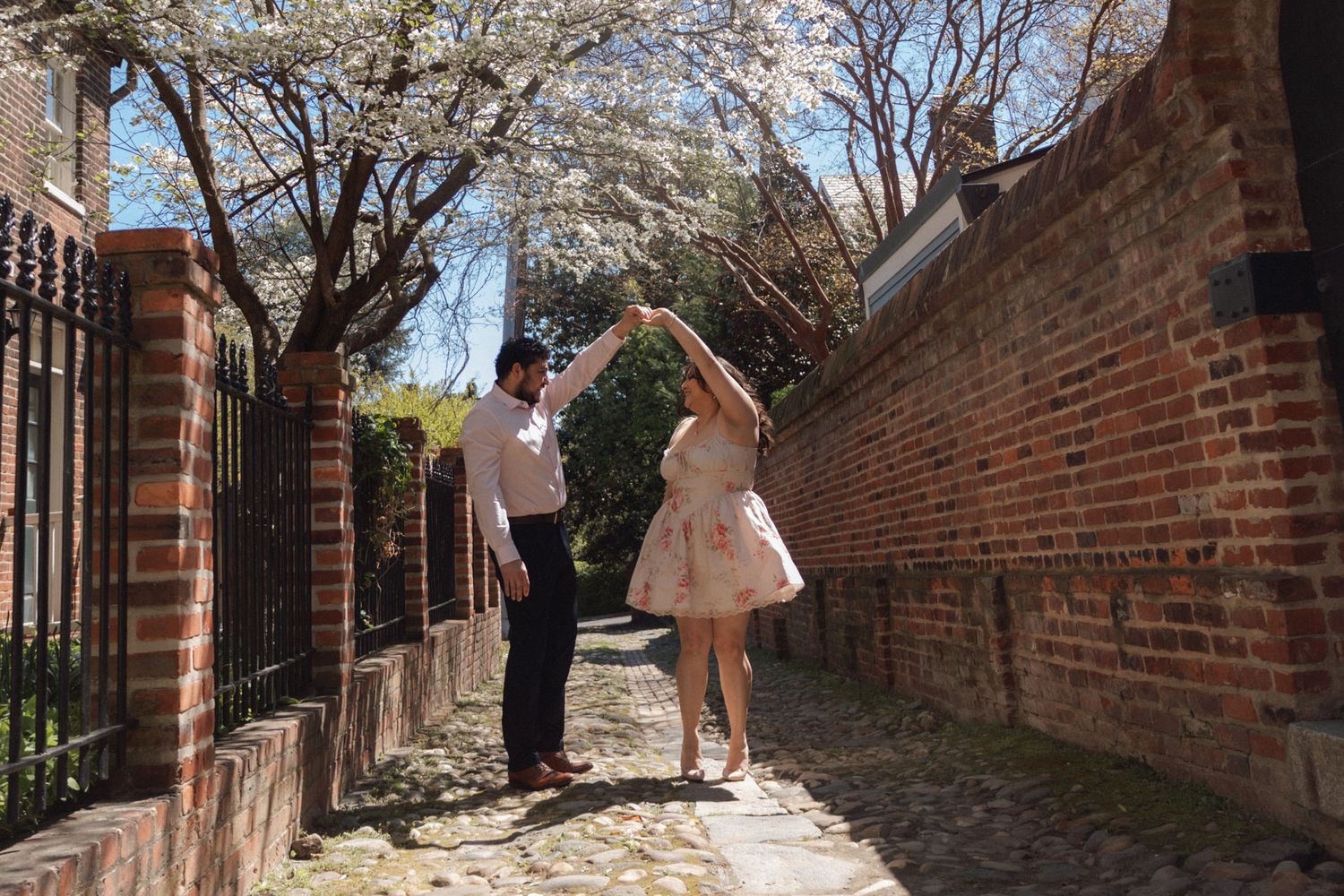 Couple dancing on a cobblestone path beside a brick wall with blooming cherry blossom trees in spring.