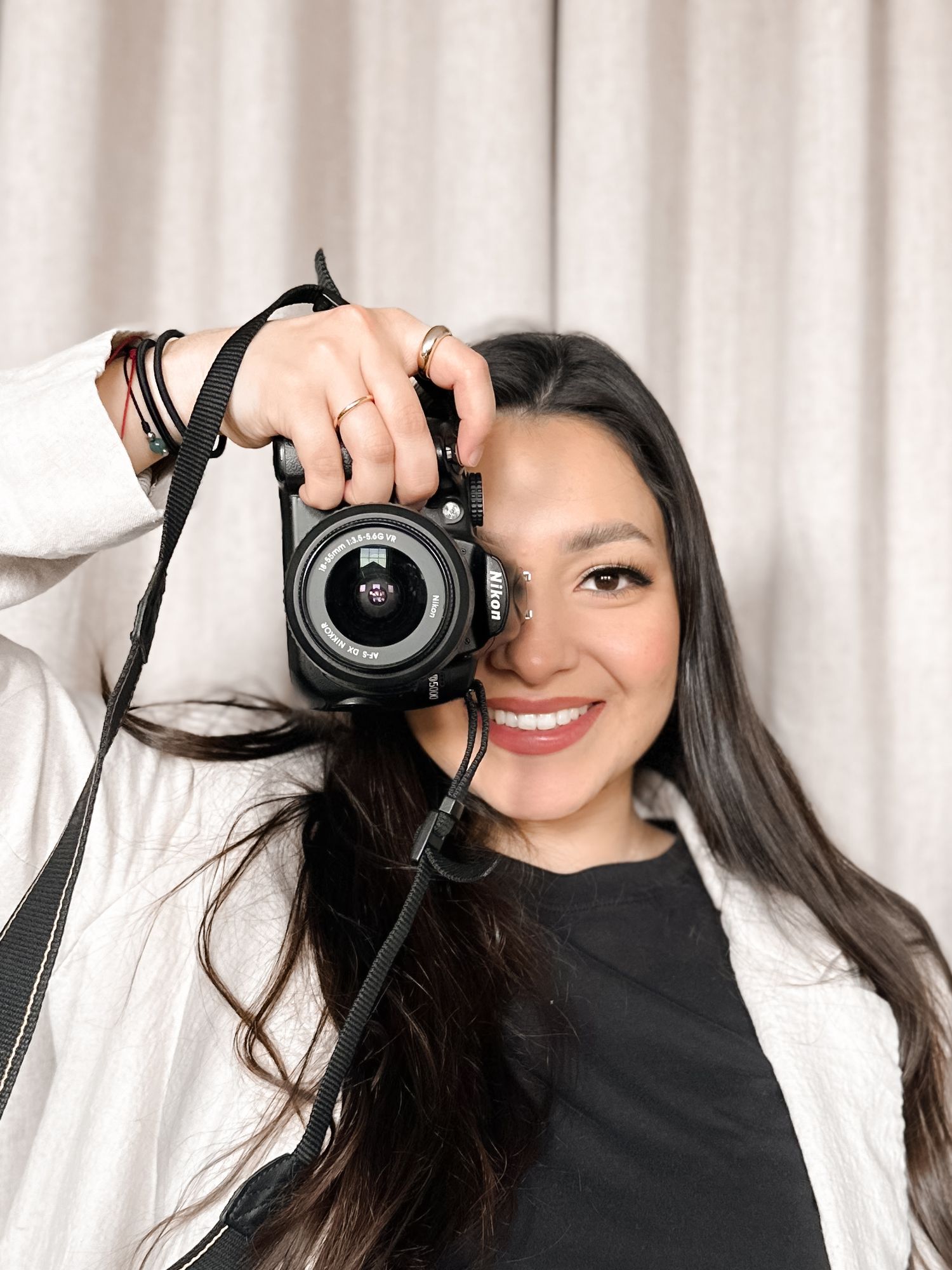 Gift of Moments owner, Tatiana, woman holding a camera up to her eye, wearing a white jacket and black shirt, with long dark hair.