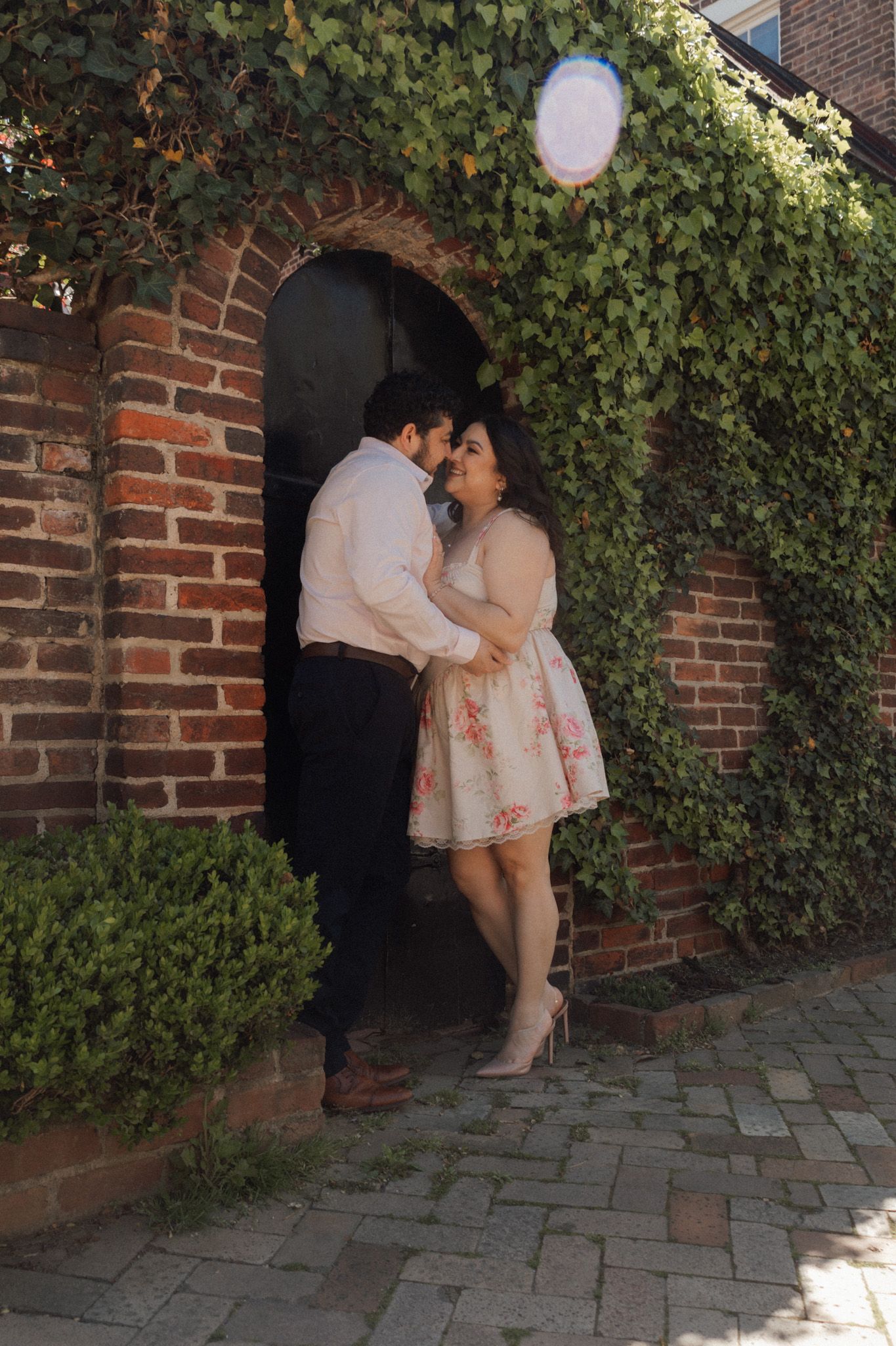 Couple embracing in arched brick doorway covered in ivy, woman in floral dress and man in white shirt.