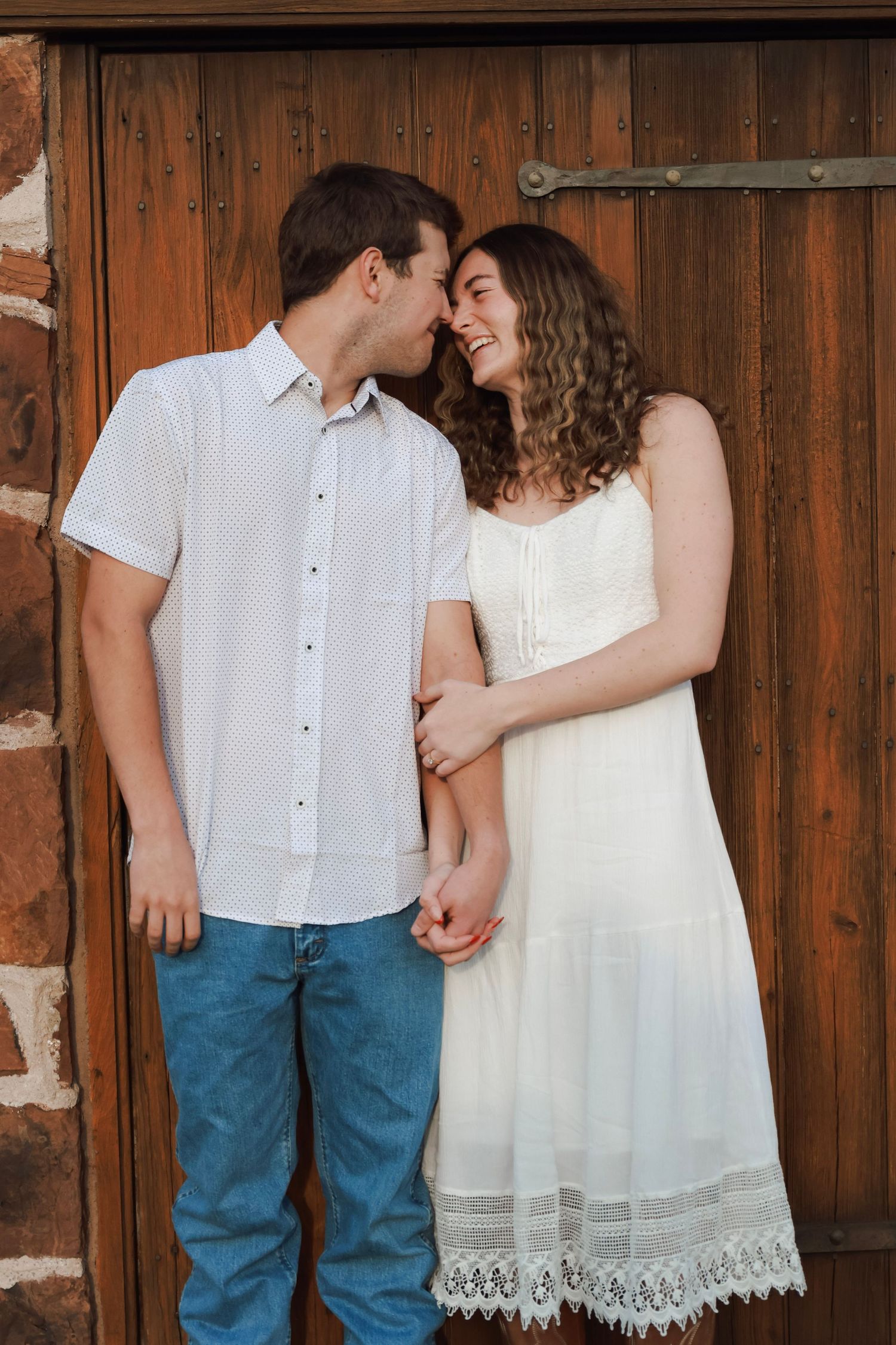 A smiling couple in white outfits shares a tender moment in front of a rustic wooden door.