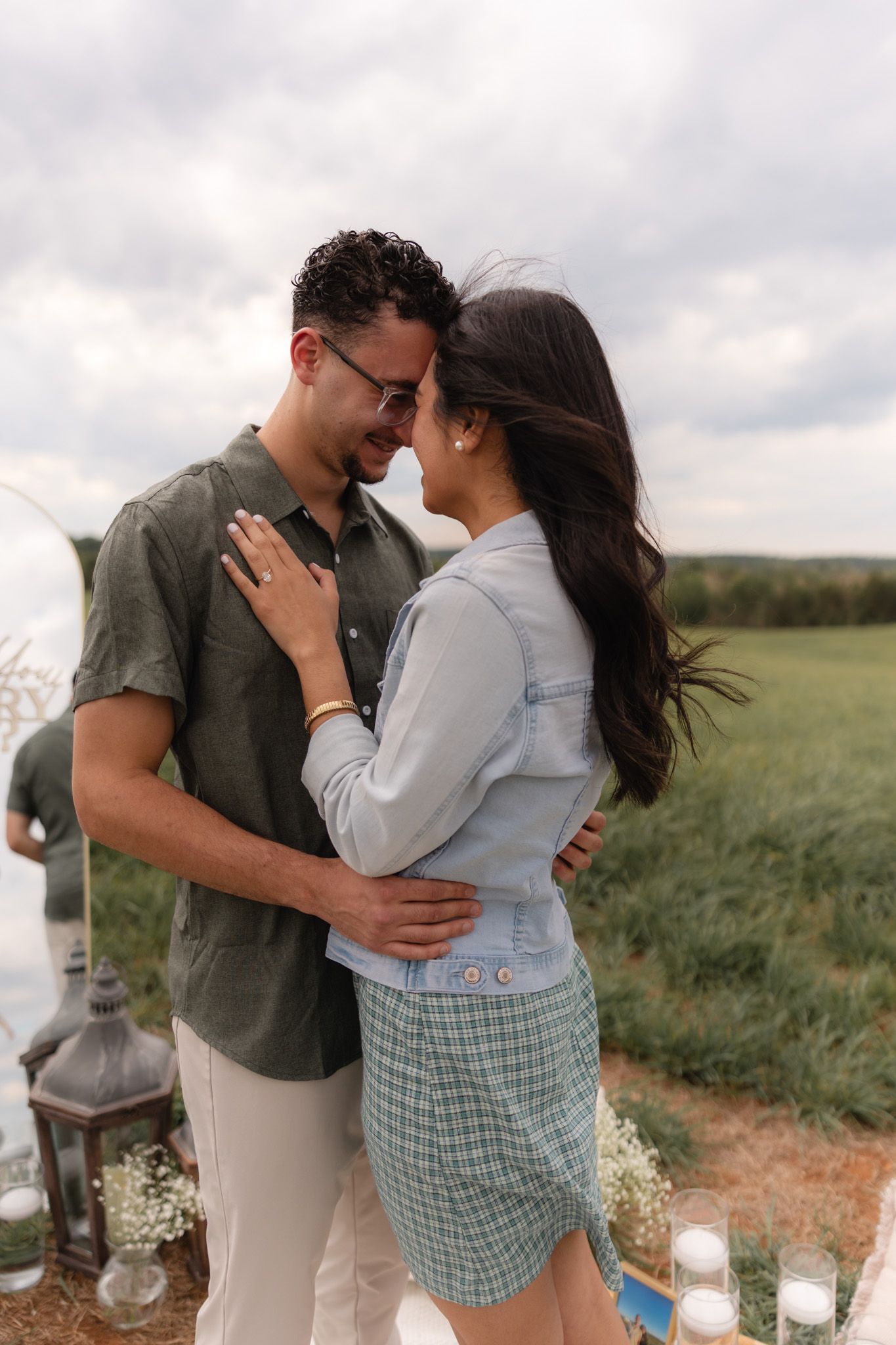 Couple sharing a romantic moment outdoors near a field, woman showing engagement ring while embracing her partner.