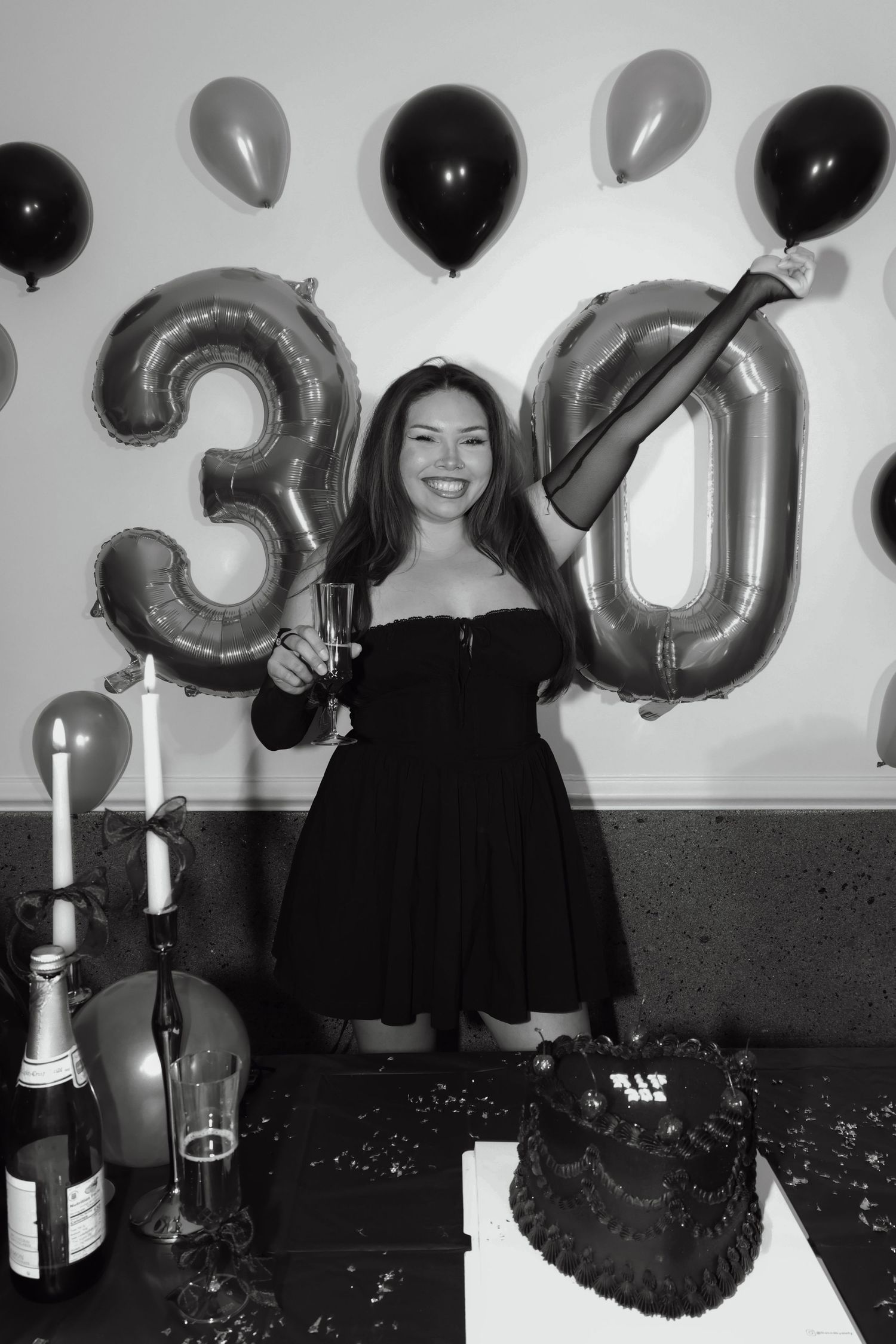 Woman in black dress celebrating 30th birthday with champagne, balloons, and cake in black and white photo.