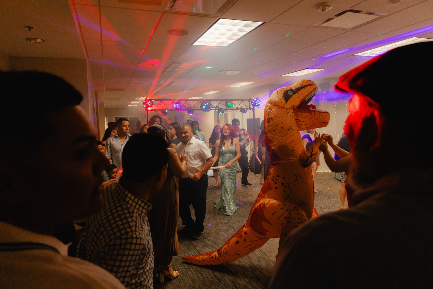 Person in inflatable T-Rex costume dancing at a party with guests in a dimly lit banquet hall.
