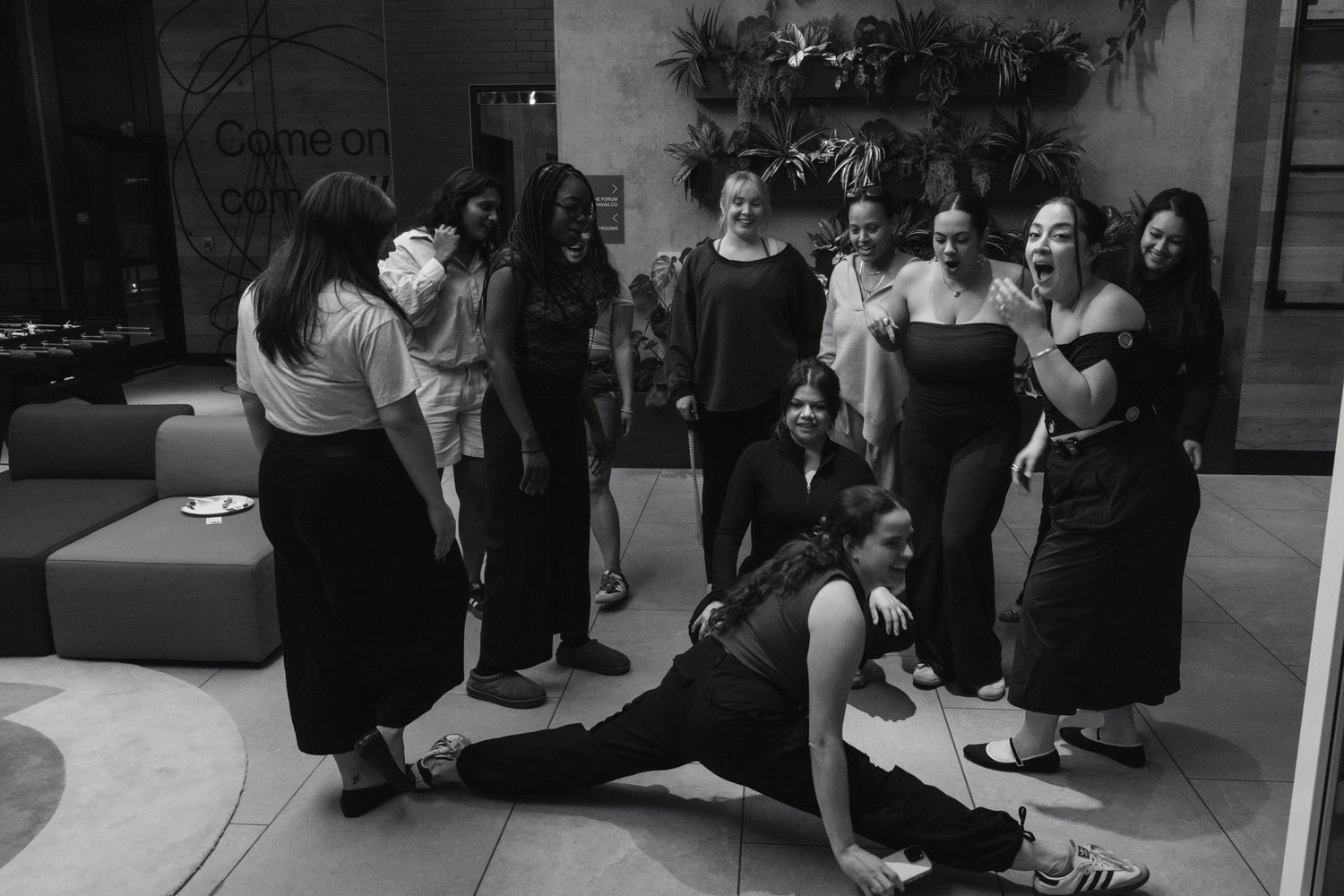 Group of women gathered in a modern lobby, one performing a dramatic floor pose in black and white photo.