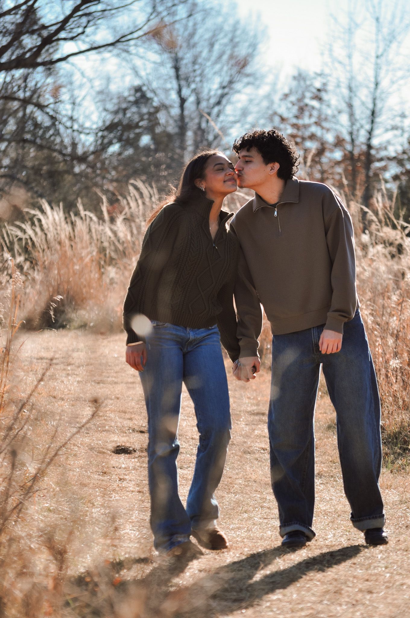 A young couple walks hand-in-hand on a rustic trail, sharing a kiss amid golden autumn grasses.