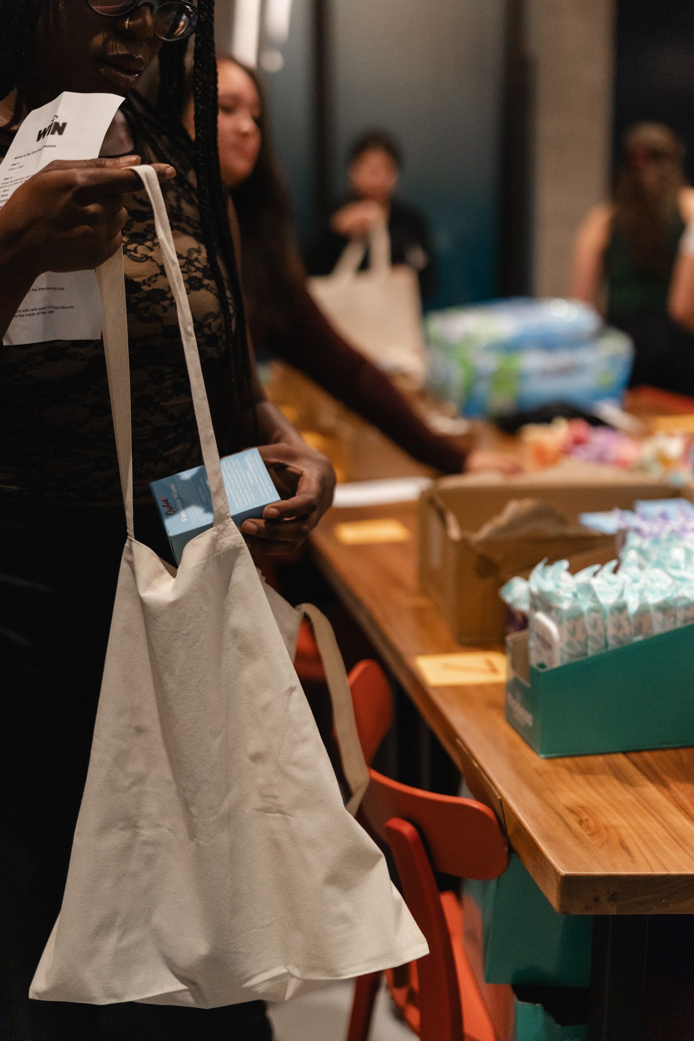 Woman holding white tote bag and small box at a community supply distribution event with diapers on table.
