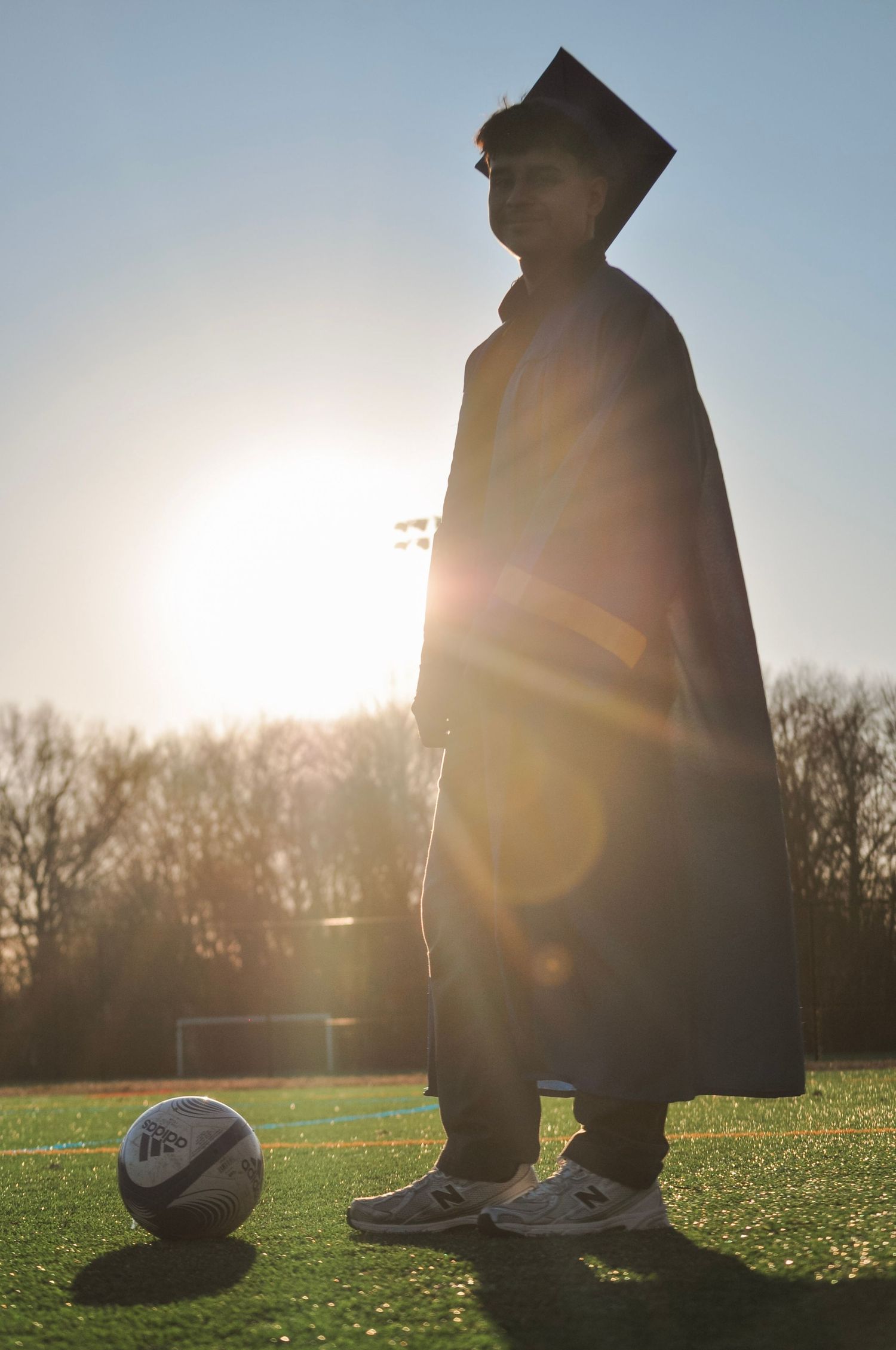 Graduate in cap and gown standing on soccer field at sunset with a soccer ball at their feet.