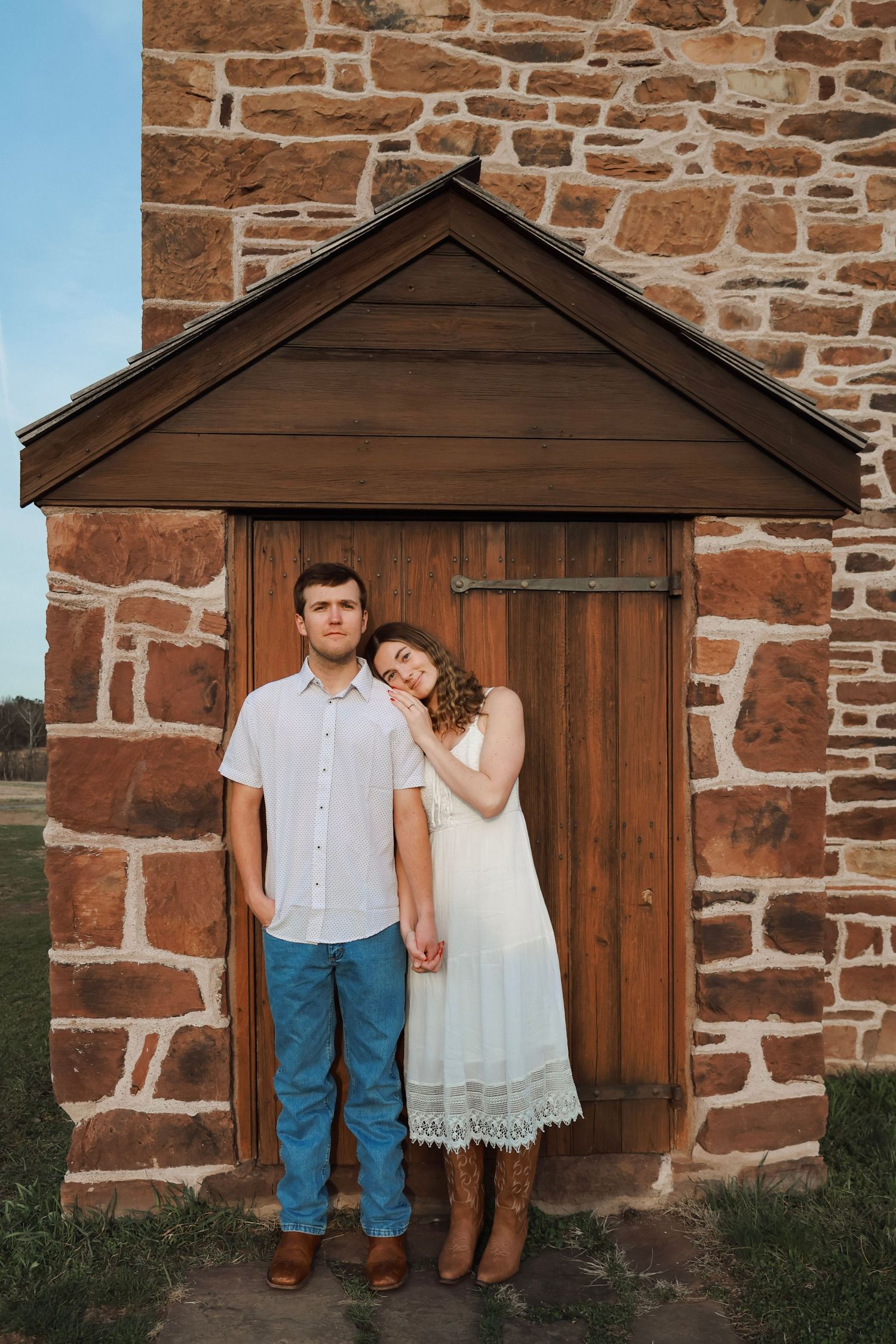 Couple posing in front of rustic wooden door and stone wall, woman in white dress leaning on man in blue jeans at national battlefield