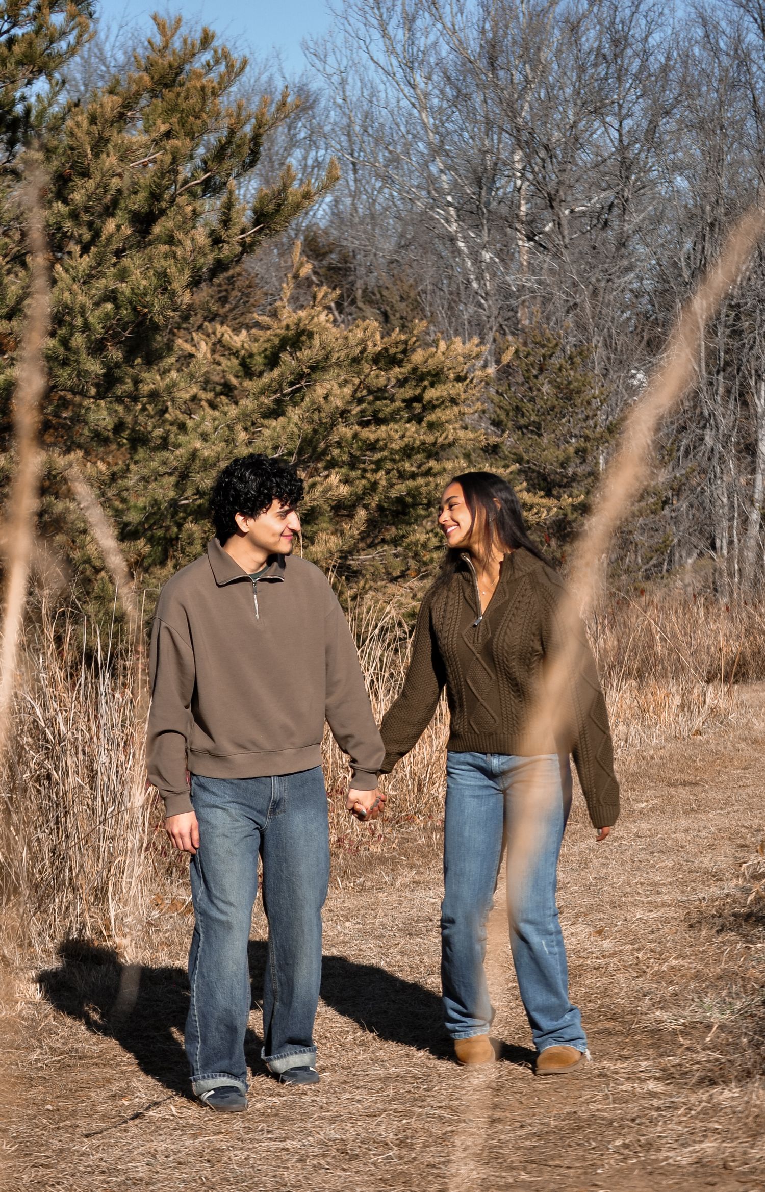 Couple holding hands and smiling at each other while walking on a nature trail surrounded by trees in autumn.