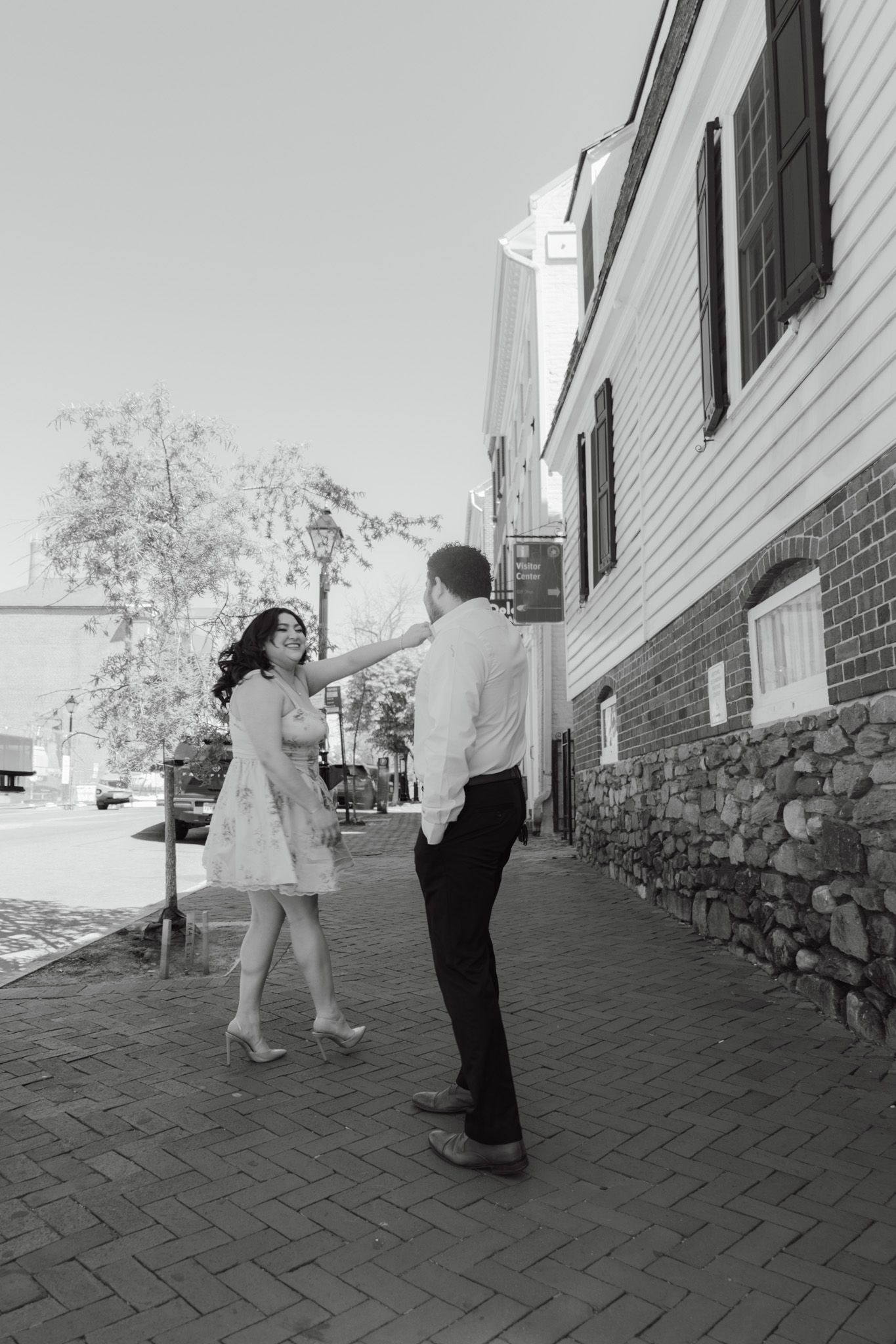 Black and white photo of a couple dancing on a brick sidewalk beside historic buildings on a sunny day.